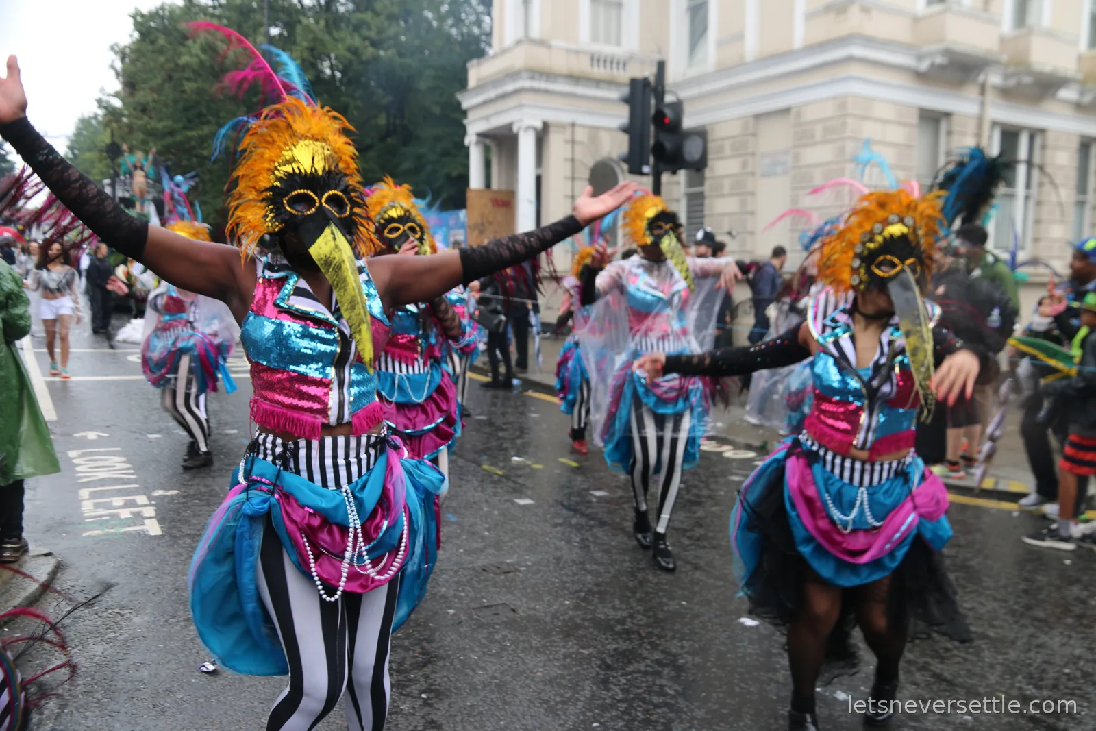 Dancers in blue and pink costumes with orange feathered headdresses and masks advancing down the road in formation at Notting Hill Carnival