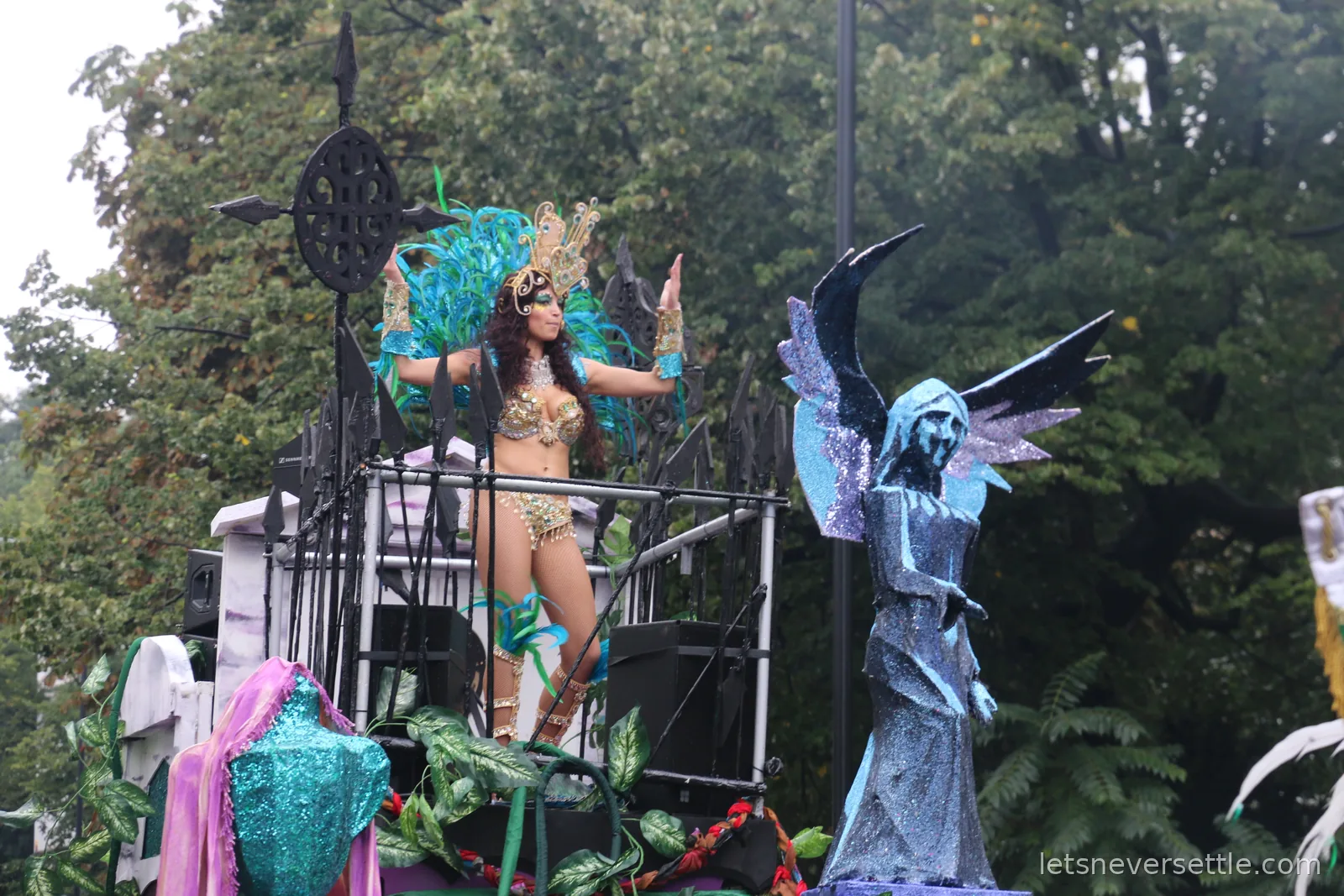 A dancer in a turquoise and gold costume performing on a carnival float beside a large blue winged angel figure, trees behind