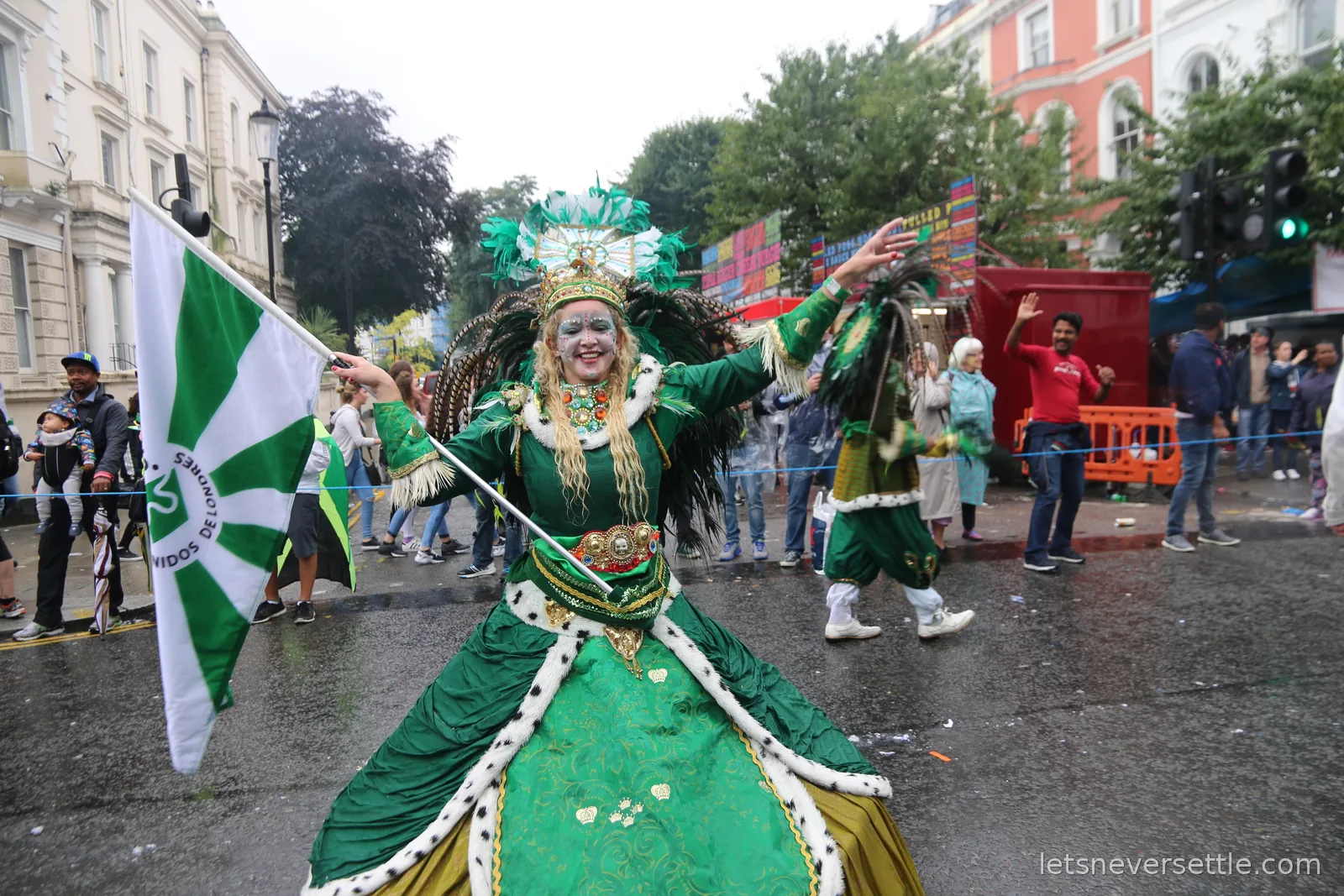 A performer in an elaborate green and gold gown waving a large green and white flag in the middle of a wet Notting Hill street