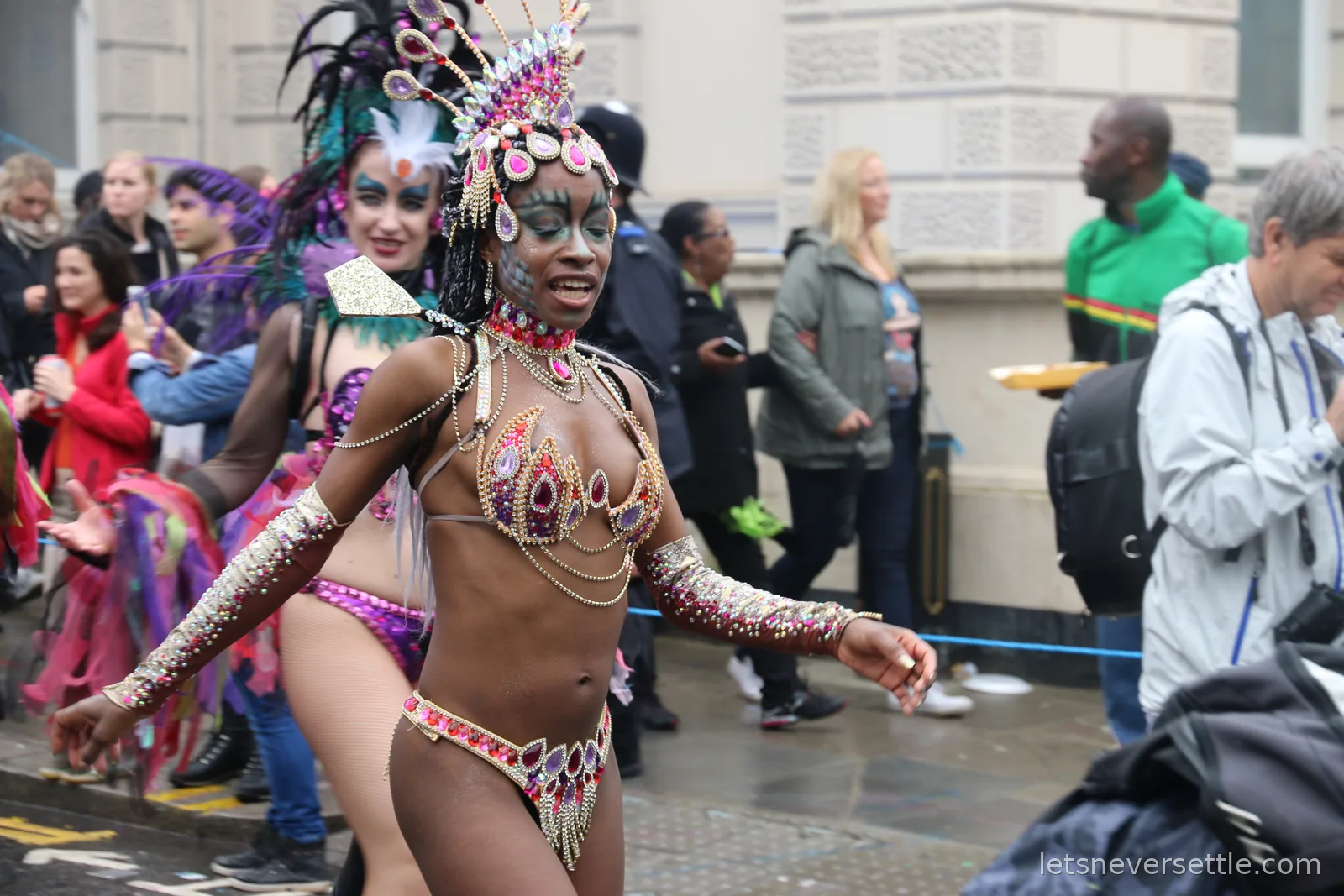 A dancer in a jewelled pink and gold costume and teardrop headdress mid-step in the Notting Hill Carnival parade