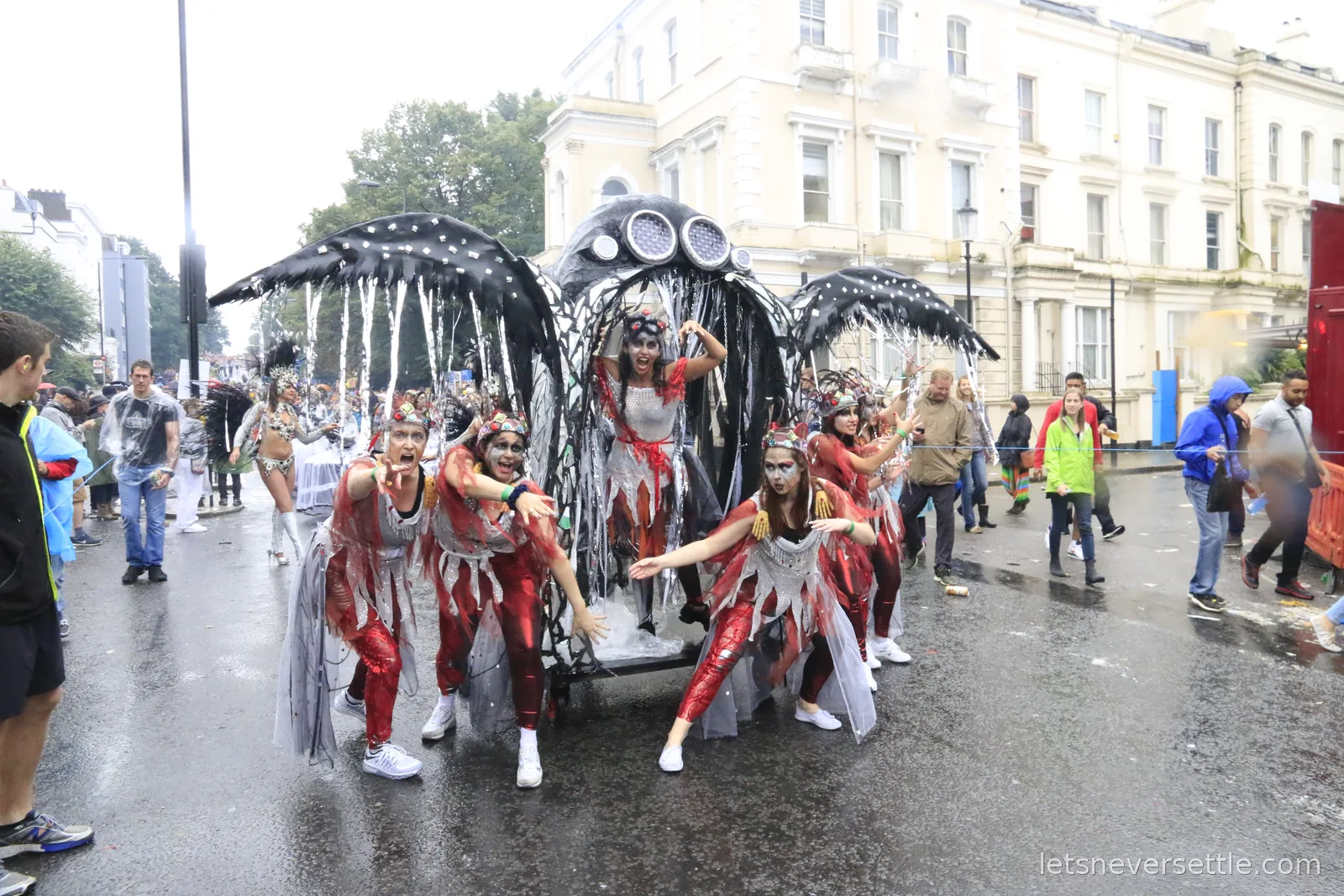 A carnival troupe in silver and red tattered costumes posing around a giant black winged creature float on a wet Notting Hill street