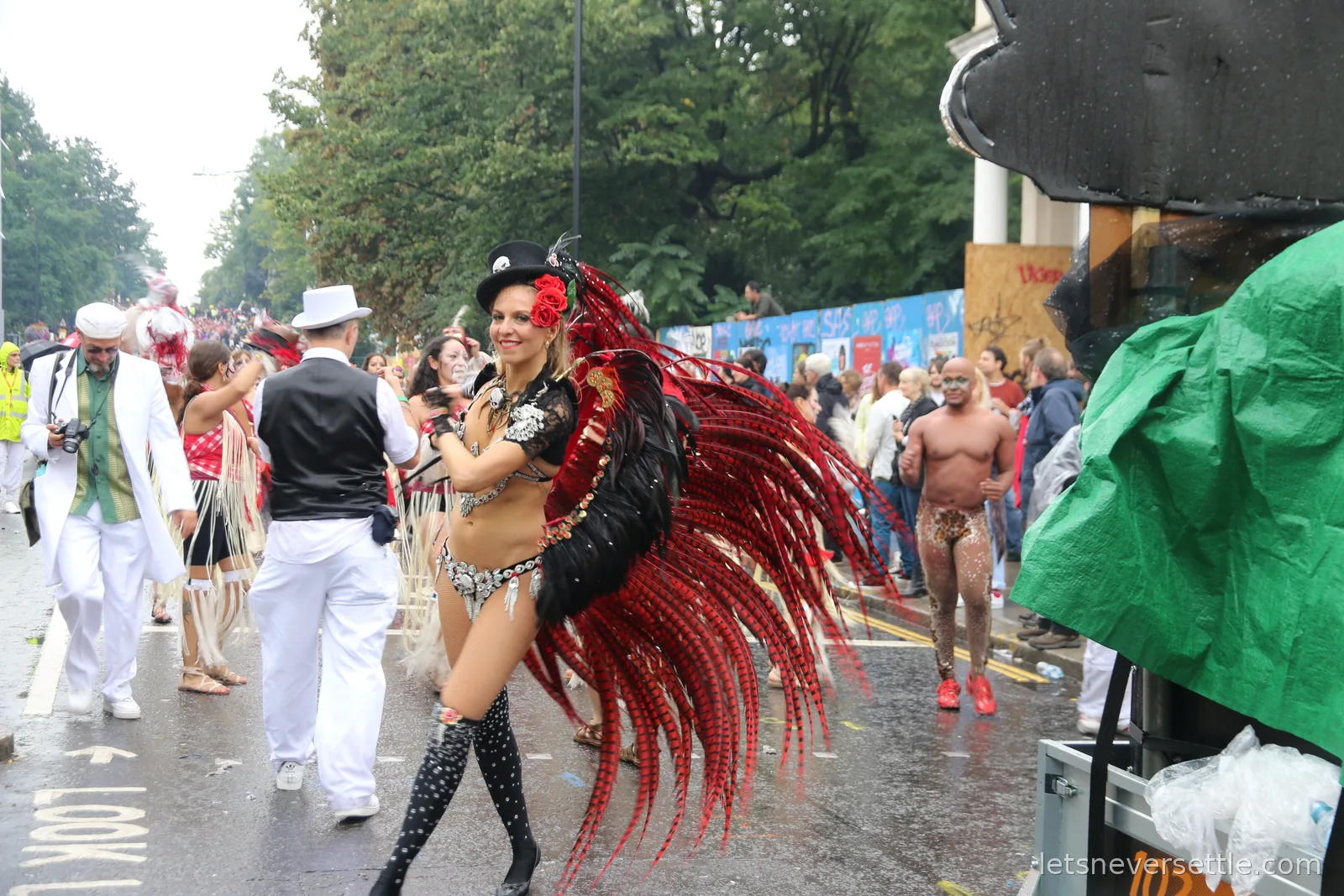 A dancer in a black costume with a huge fan of red and black feathers walking up the wet street past band members dressed in white