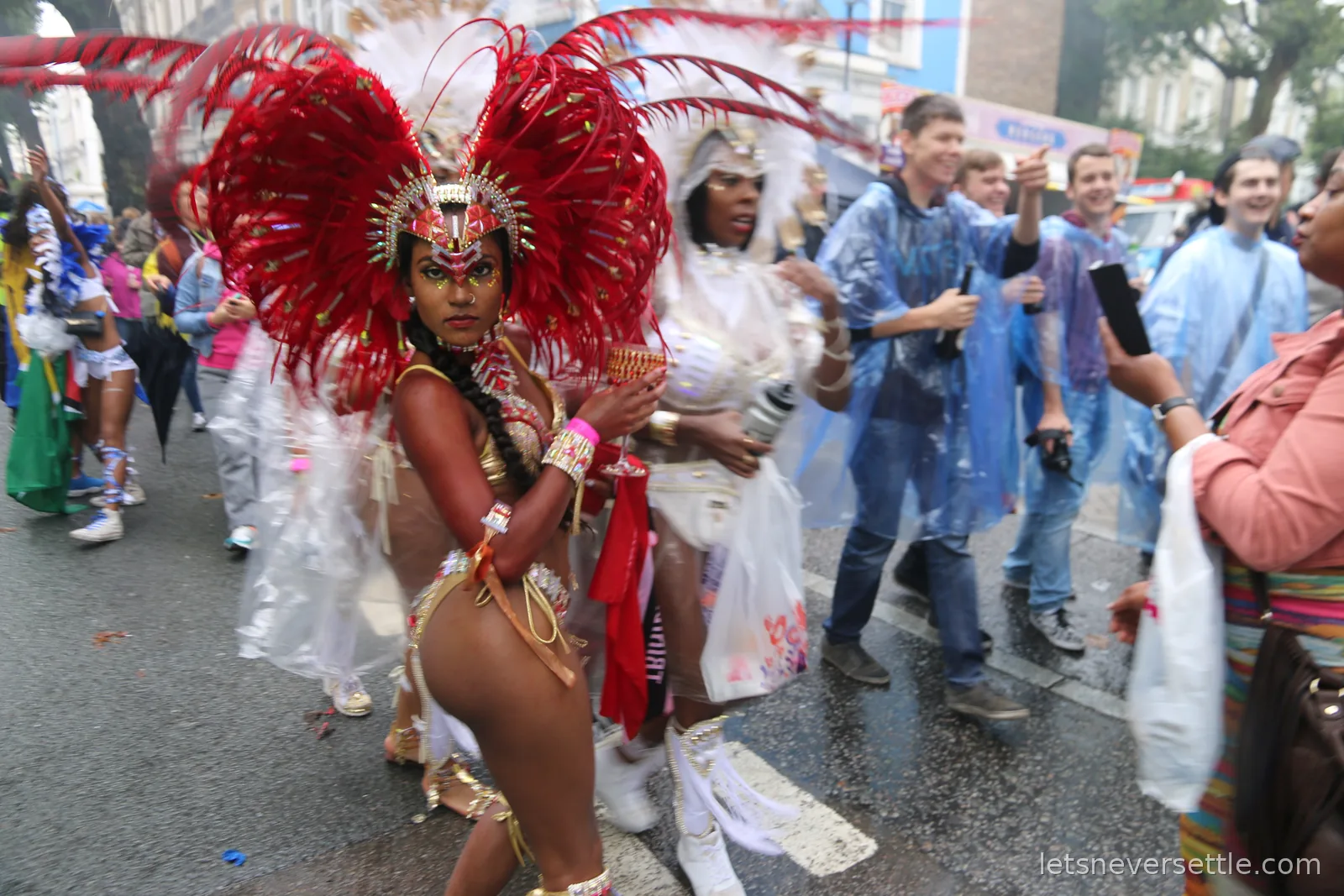 A carnival dancer in a towering red feathered headdress and gold costume leading the parade past spectators in rain ponchos at Notting Hill Carnival