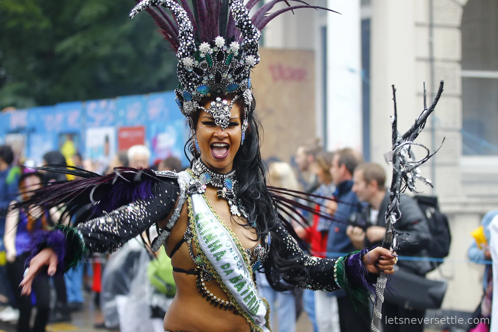 A dancer in a jewelled costume and tall purple and silver headdress smiling mid-step in the parade, spectators close behind her