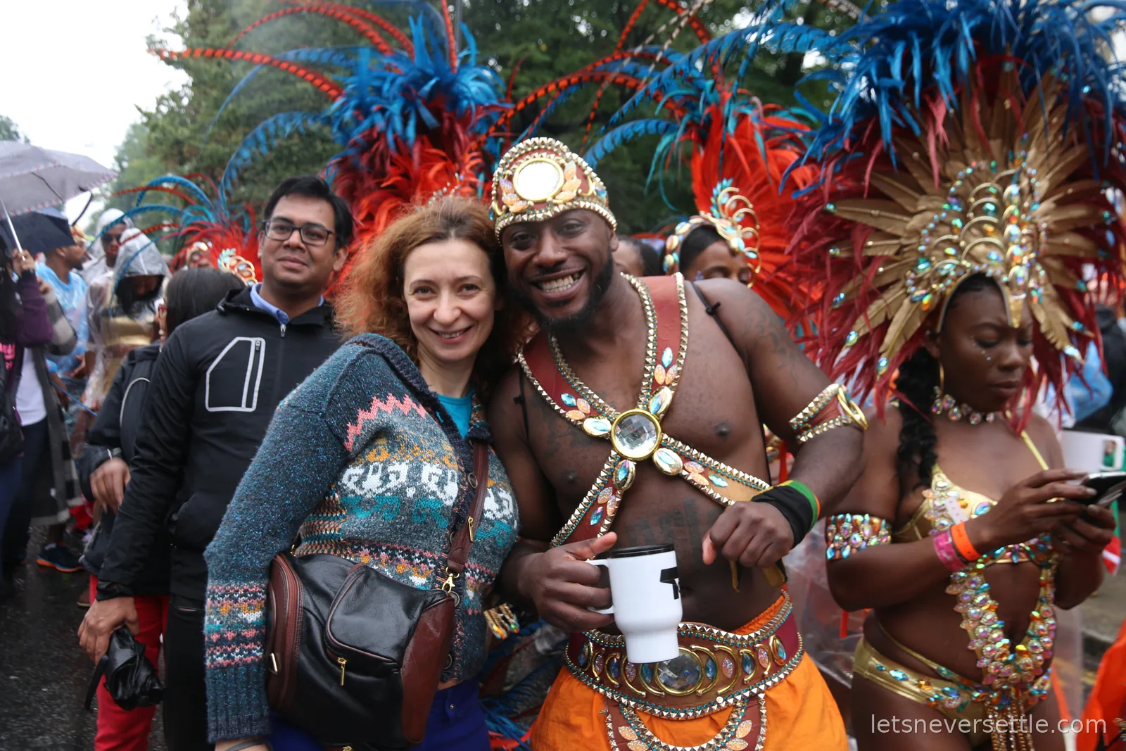 Inga and Dileep in coats posing with two carnival performers in red and blue feathered headdresses and jewelled costumes on a wet street