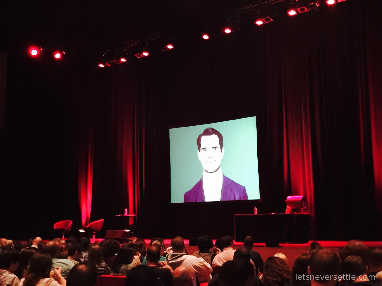 A dark auditorium lit red from above, the stage bare except for two chairs and a table, and a big screen showing a stylised portrait of comedian Jimmy Carr in a purple jacket