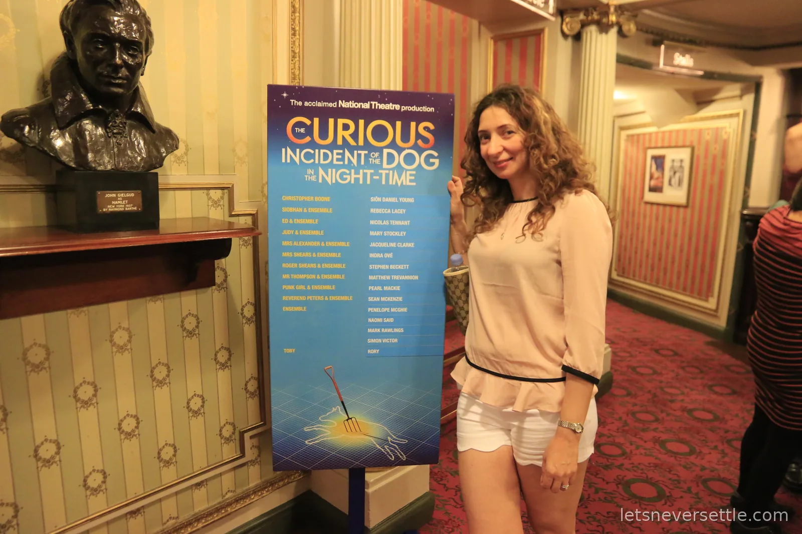 Inga beside a poster for the National Theatre production of The Curious Incident of the Dog in the Night-Time, with a bust of John Gielgud on a shelf nearby