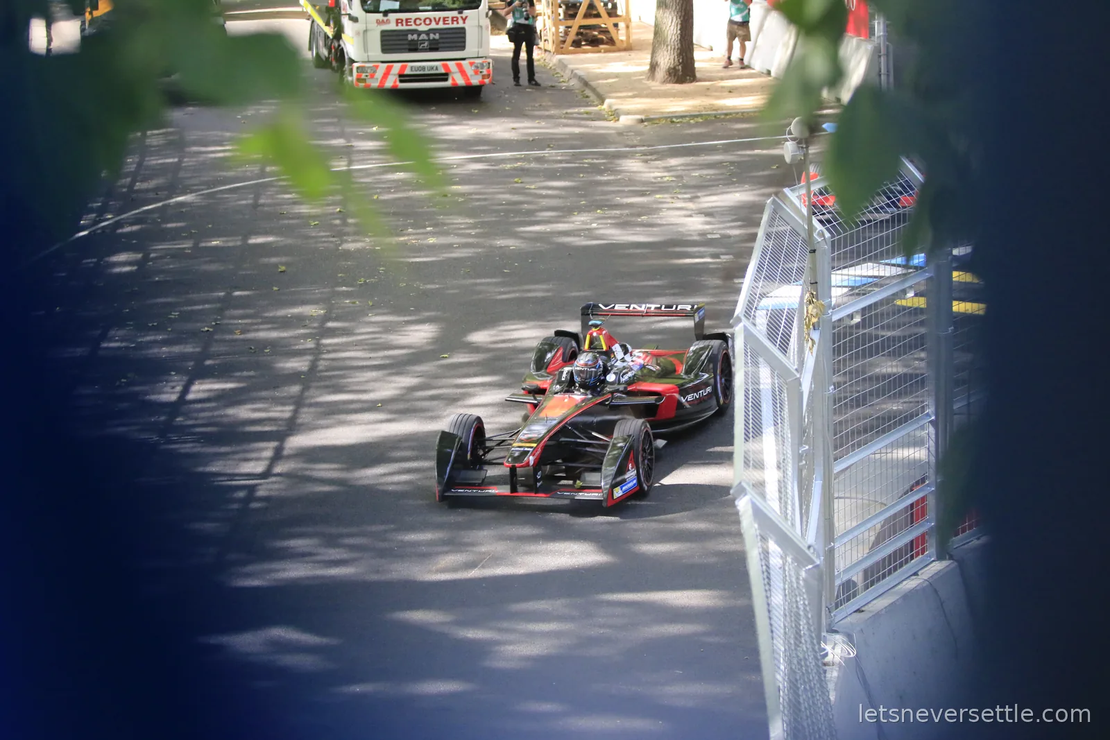 A Formula E car on the circuit seen through overhanging leaves, with catch fencing and a recovery truck along the track edge in Battersea Park