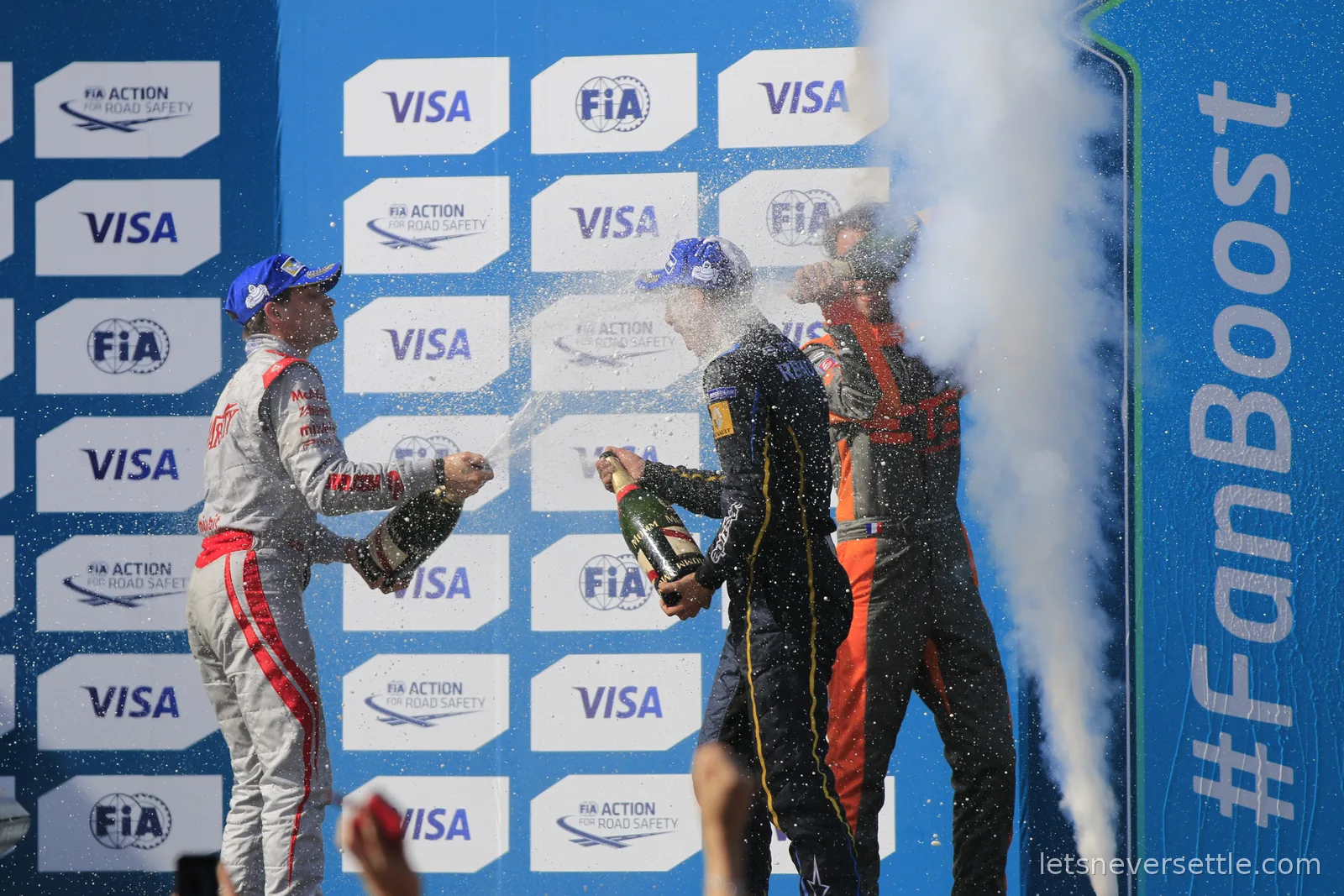 Three drivers in race suits spraying champagne on the podium in front of a Visa and FIA branded wall