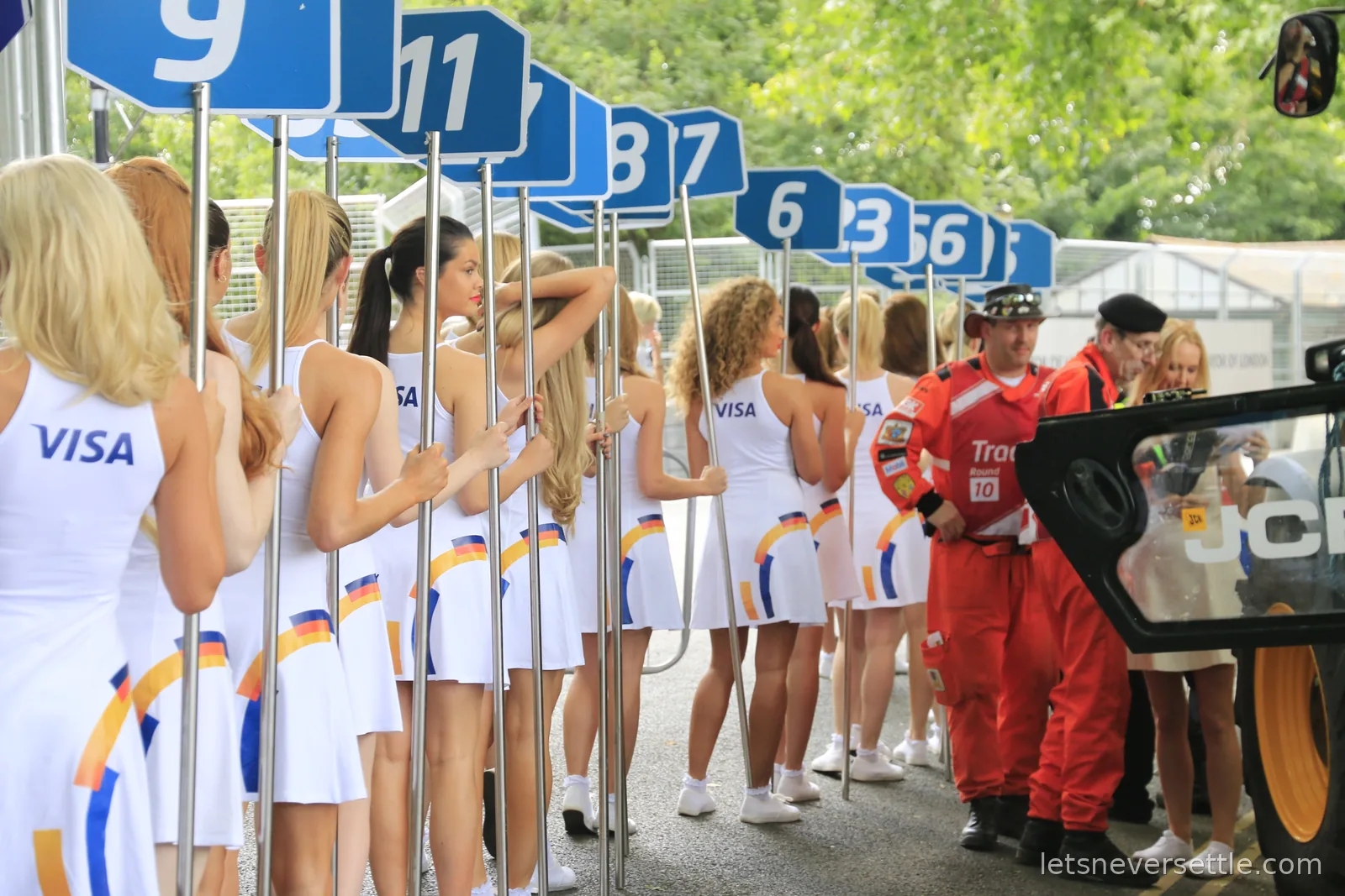 A line of grid hosts in white Visa dresses holding numbered blue grid boards, with marshals in red overalls standing alongside