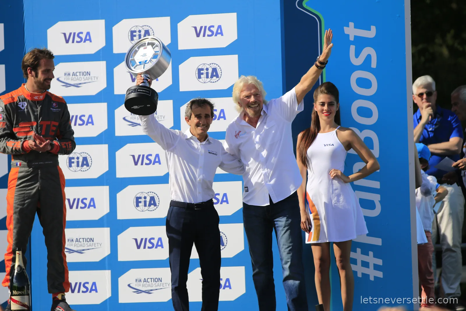Alain Prost raising a trophy beside Richard Branson with his arms spread wide on the Formula E podium