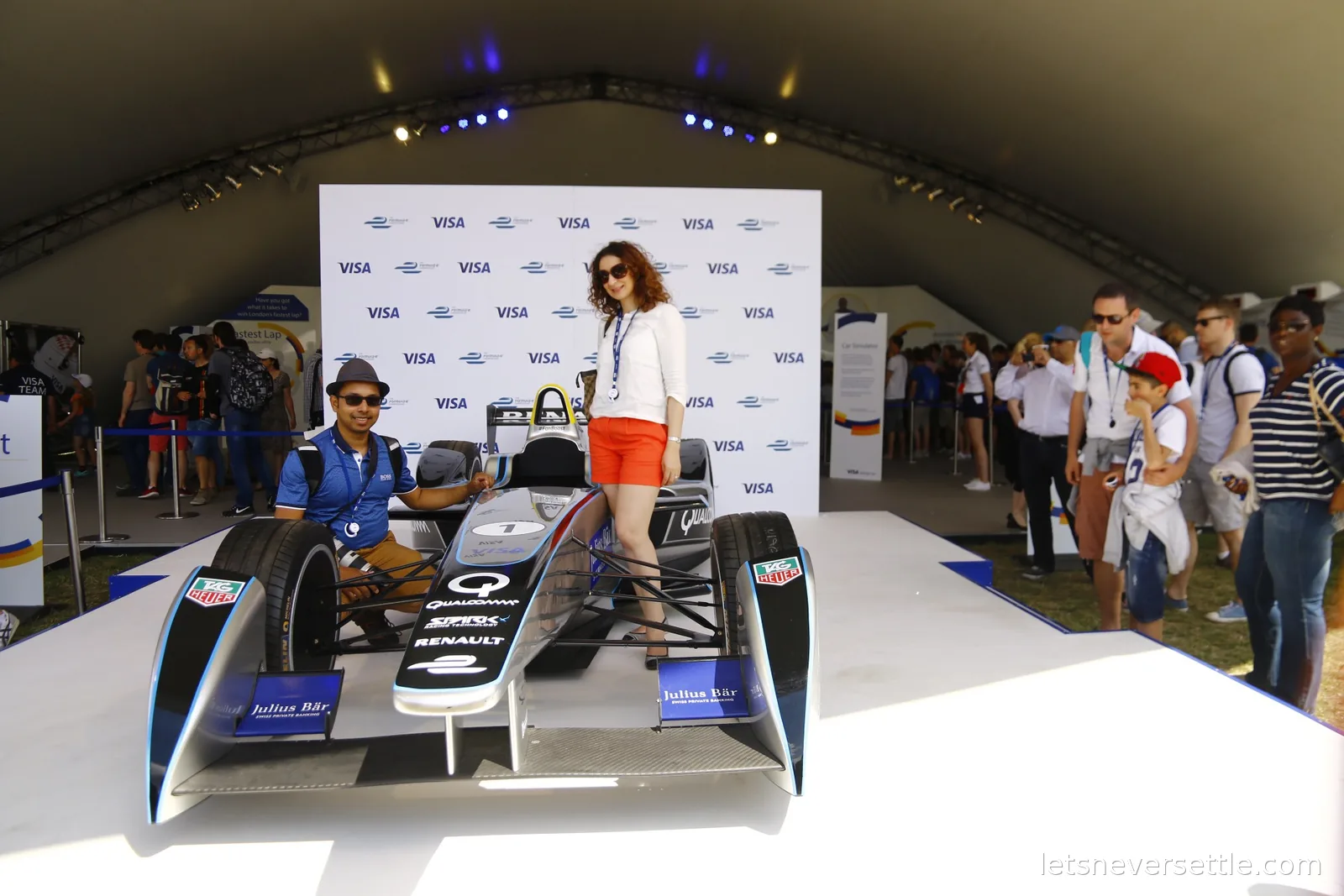 Dileep crouching and Inga standing beside a Formula E show car on a white display plinth inside the fan village tent