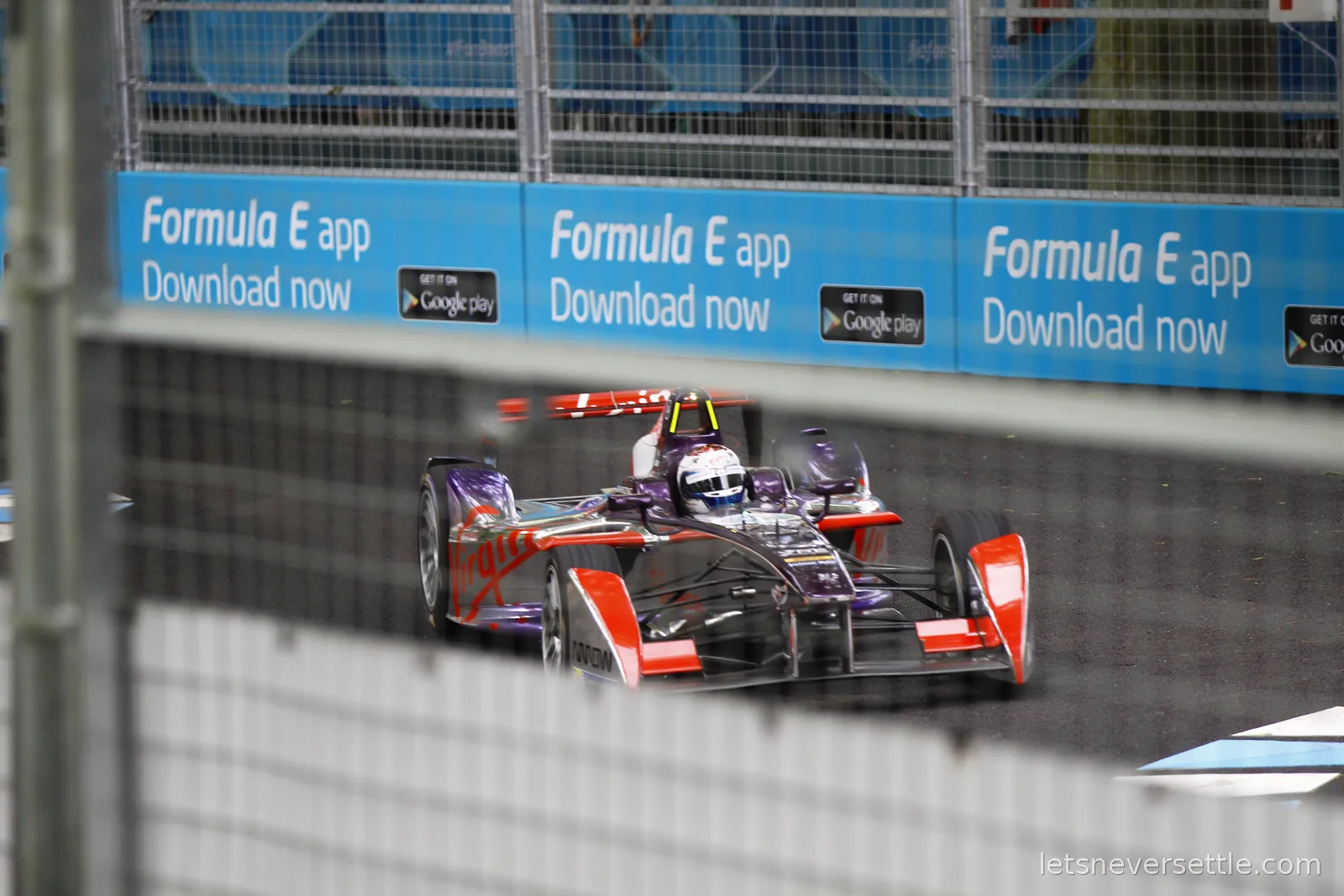 A purple and red Virgin-liveried Formula E car at speed behind catch fencing lined with Formula E app banners