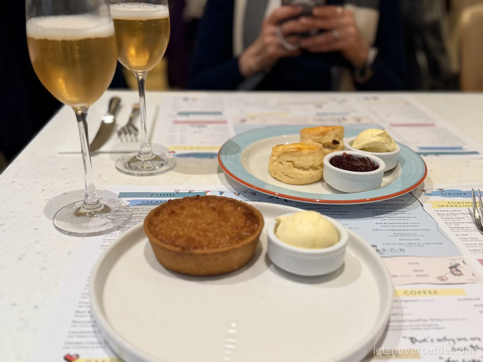 Close view of two scones with jam and clotted cream, a treacle tart and two glasses of sparkling wine on a Fortnum and Mason printed menu placemat
