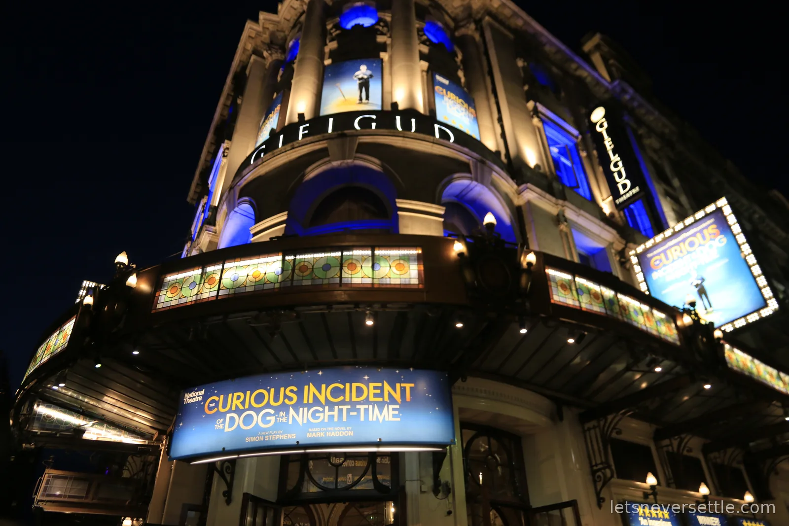 The Gielgud Theatre lit up blue at night, its curved facade carrying signage for The Curious Incident of the Dog in the Night-Time