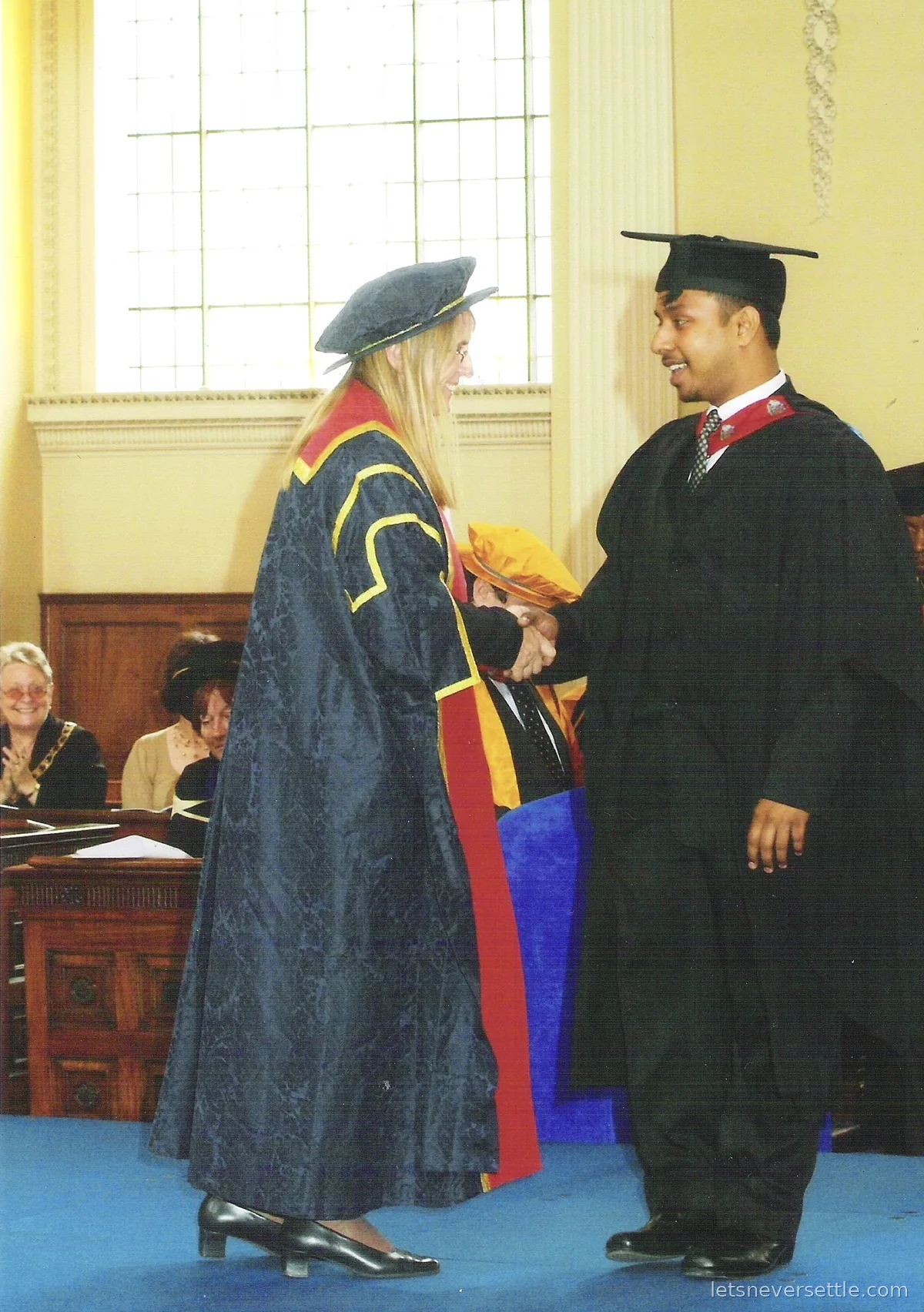 Dileep in cap and gown shaking hands on stage with an academic in a dark blue and red ceremonial robe
