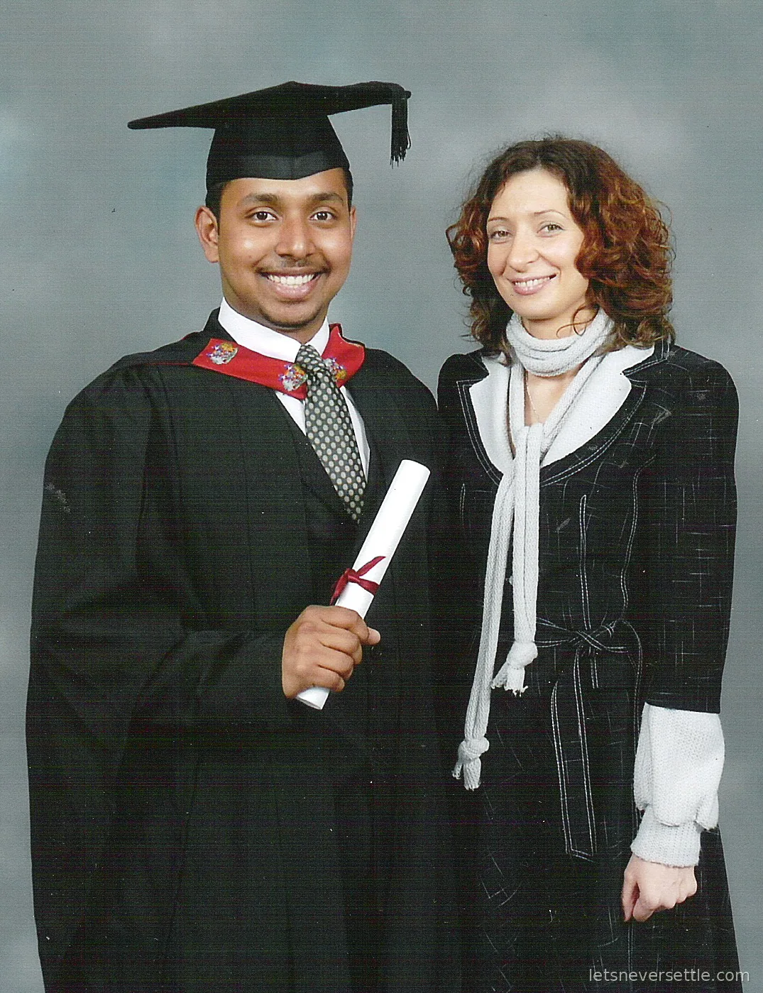 Dileep in cap and gown with his scroll standing beside Inga in a black coat and white scarf for a studio portrait