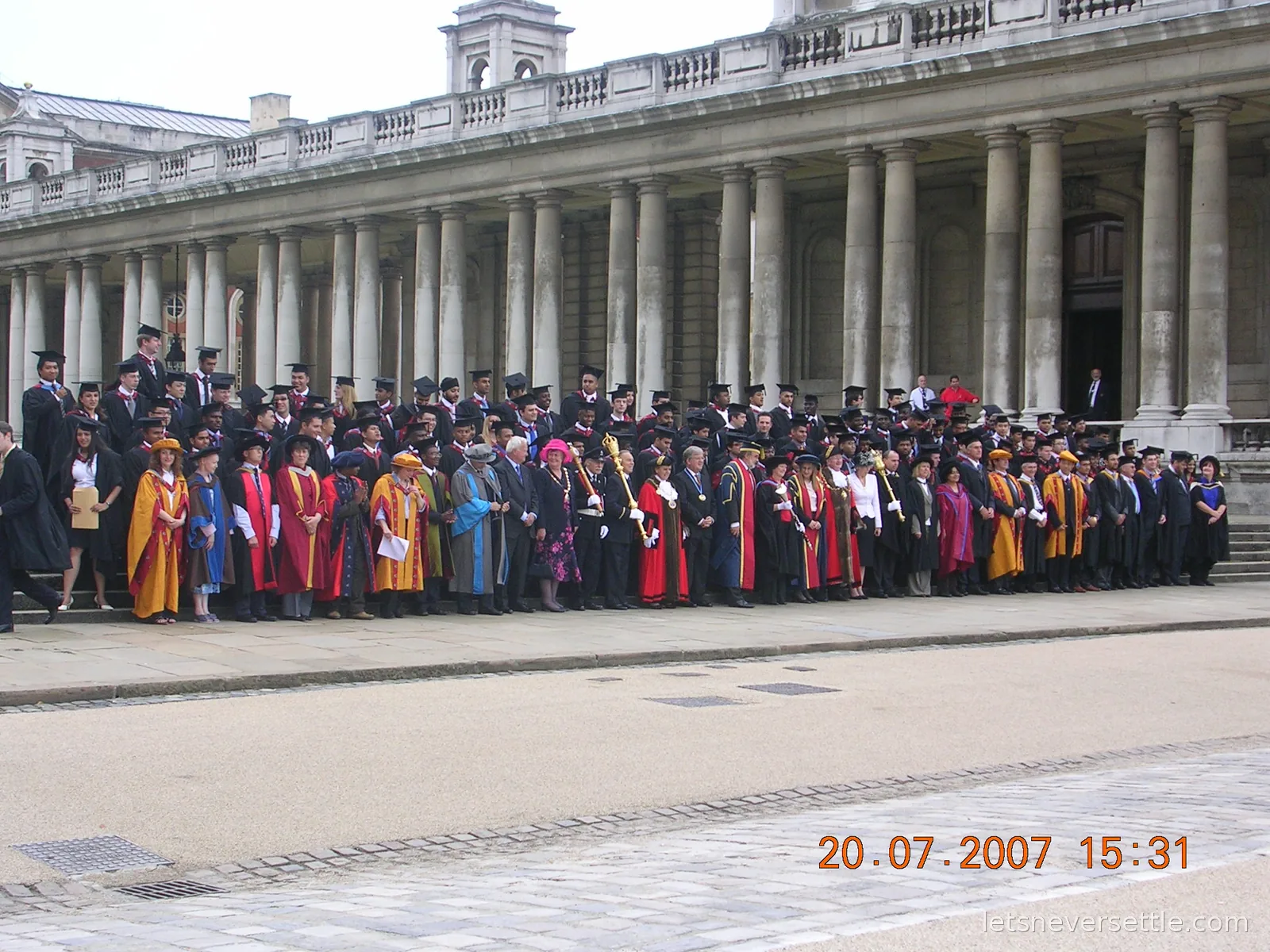 Graduates in gowns and mortarboards lined up with academics in colourful robes along the stone colonnade of the Old Royal Naval College in Greenwich