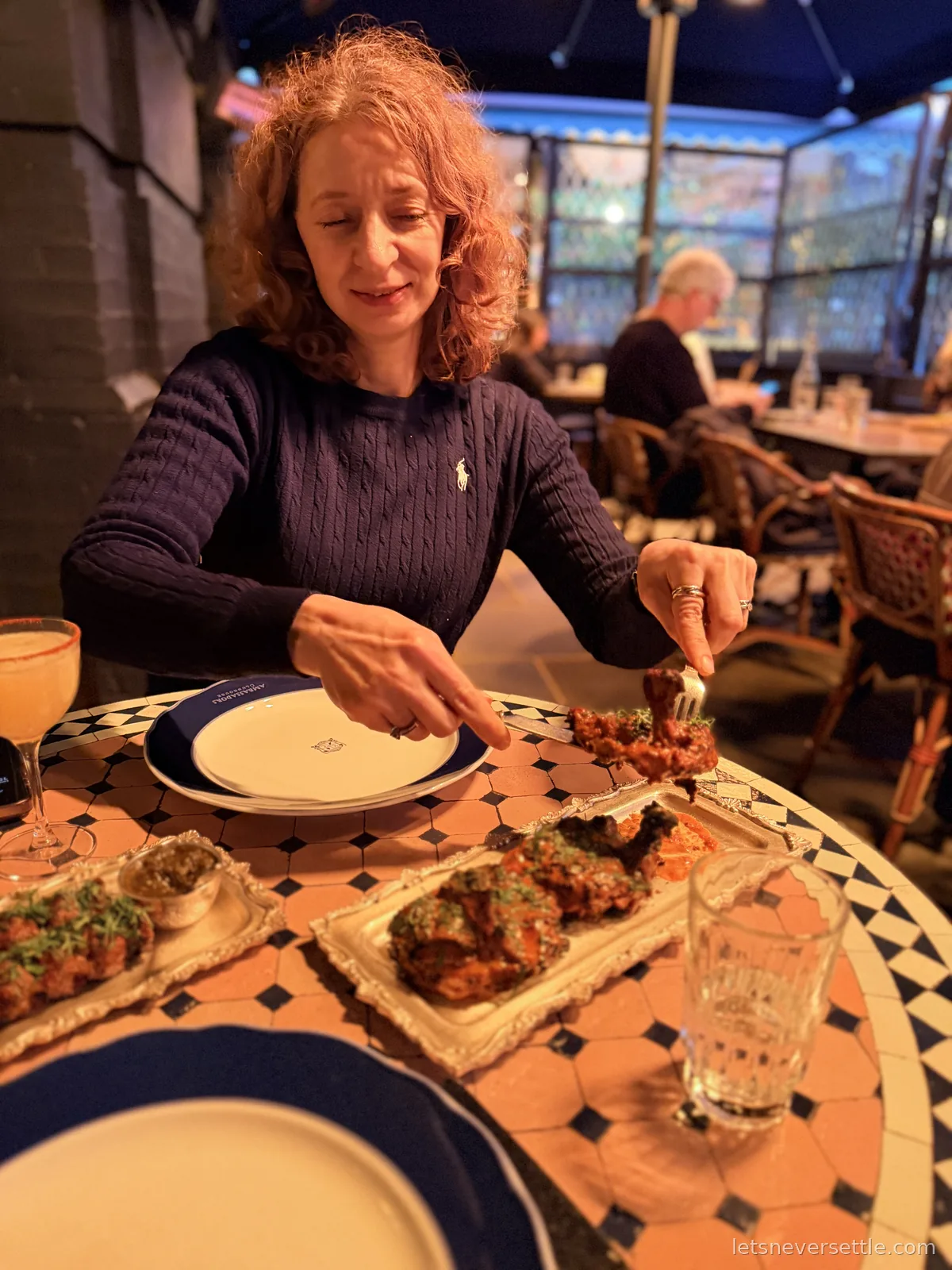 Inga lifting a grilled skewer off an ornate silver platter at a mosaic tiled table under a heated awning, more skewers, chutneys and a cocktail spread across the table