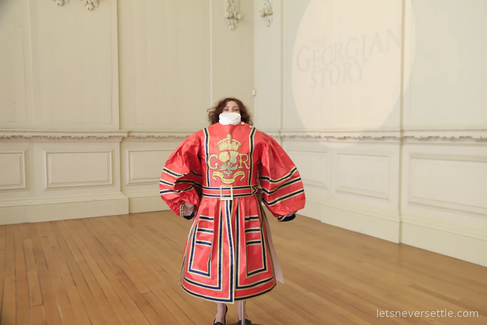 Inga wearing a red royal livery coat with a gold crown and Tudor rose badge and a white ruff, standing in a panelled room signed The Georgian Story