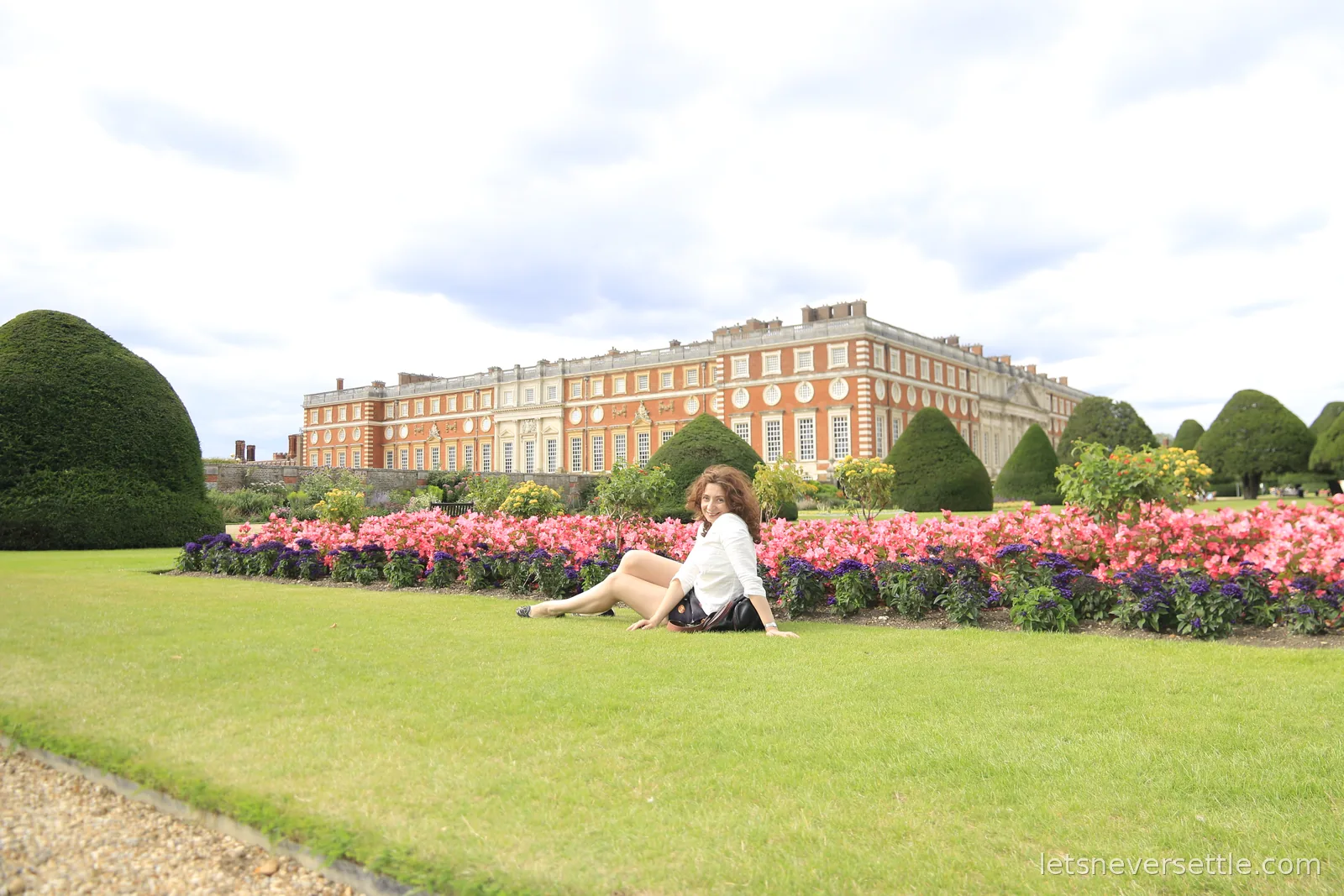 Inga sitting on the lawn in front of the baroque east front of Hampton Court Palace, pink flower beds and clipped topiary behind