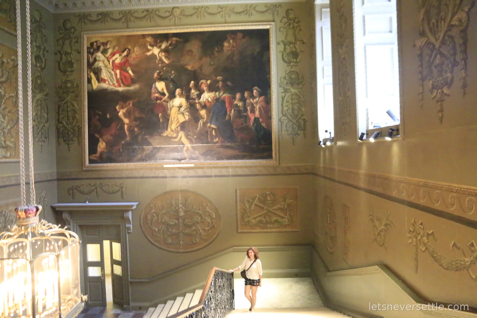 A grand painted stairwell at Hampton Court Palace with an enormous mythological canvas above moulded walls and a lantern topped with a gold crown, Inga at the foot of the stairs