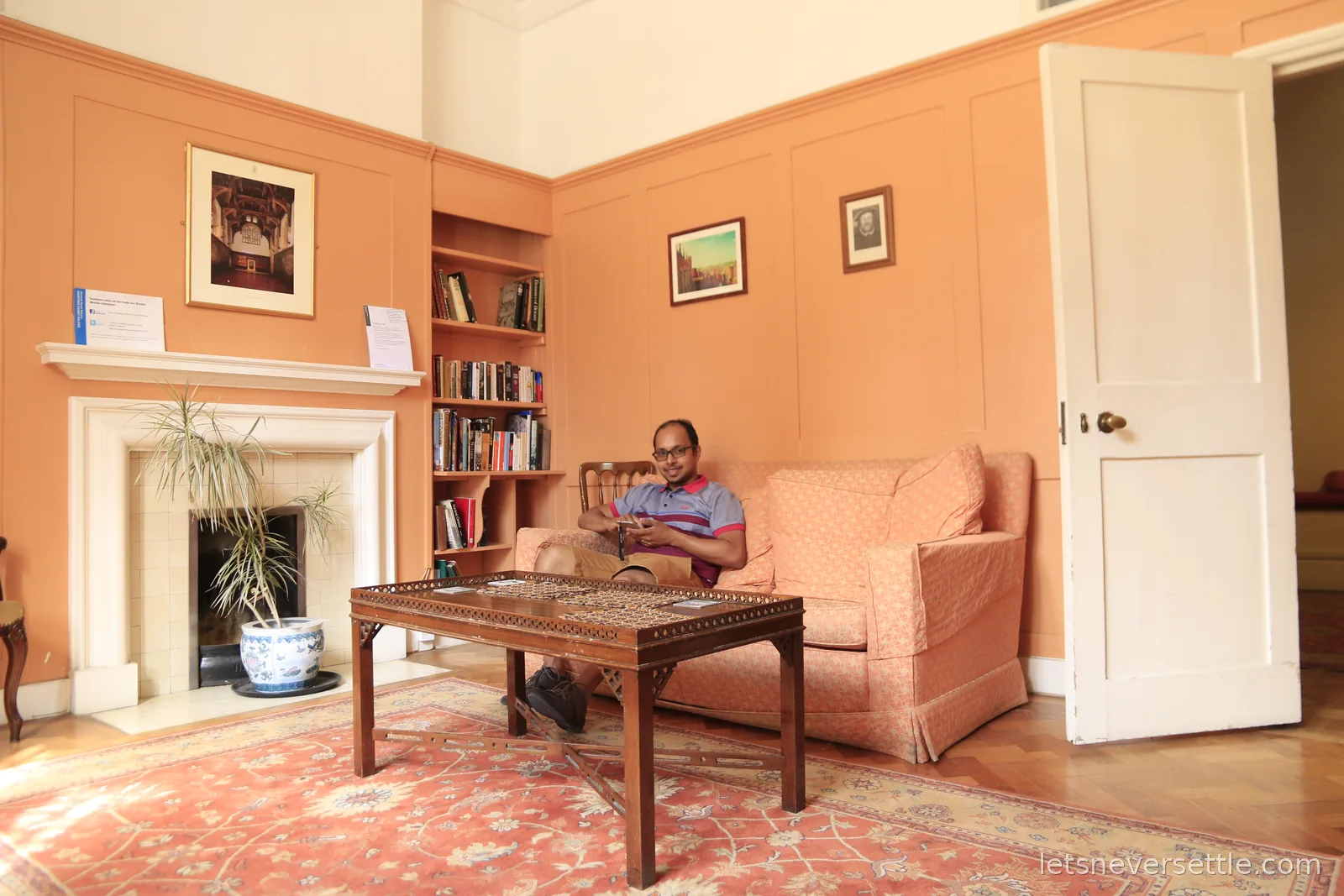 Dileep sitting in an armchair in the peach-walled members' room at Hampton Court Palace, with a fireplace, bookshelves, a low table and a patterned rug
