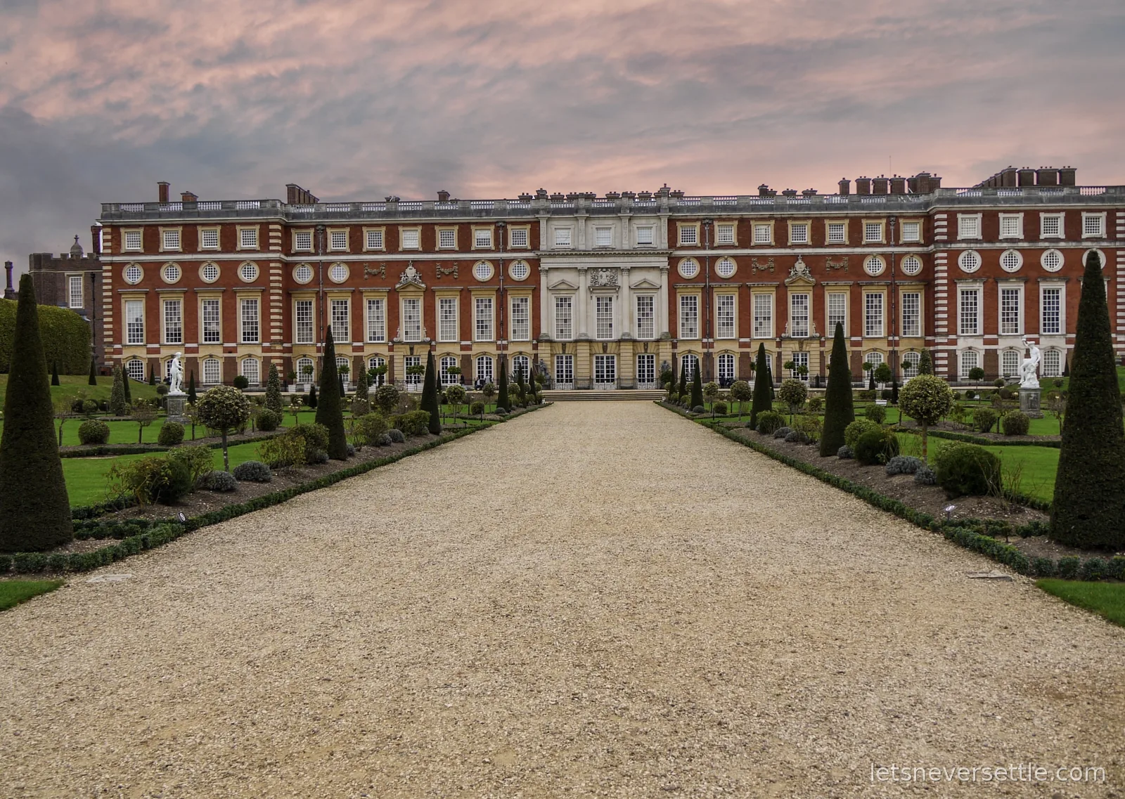 The palace front at Hampton Court under a pink evening sky, with clipped conical yews lining the gravel walk