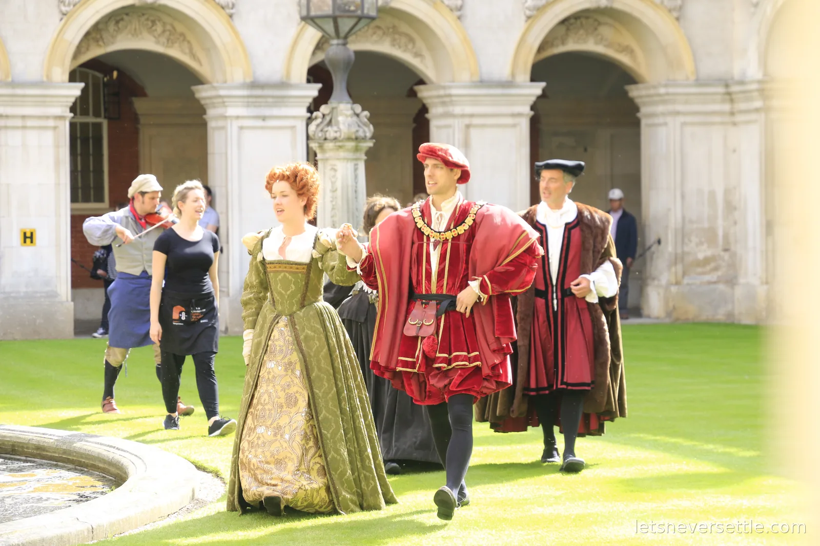 Costumed Tudor courtiers walking through a cobbled courtyard at Hampton Court Palace led by a fiddler