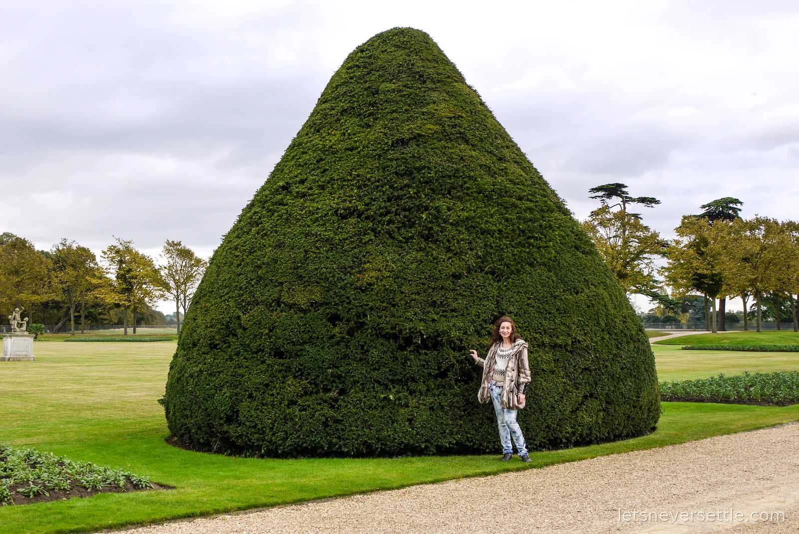 Inga standing at the base of a giant cone-clipped yew tree in the gardens of Hampton Court Palace, for scale
