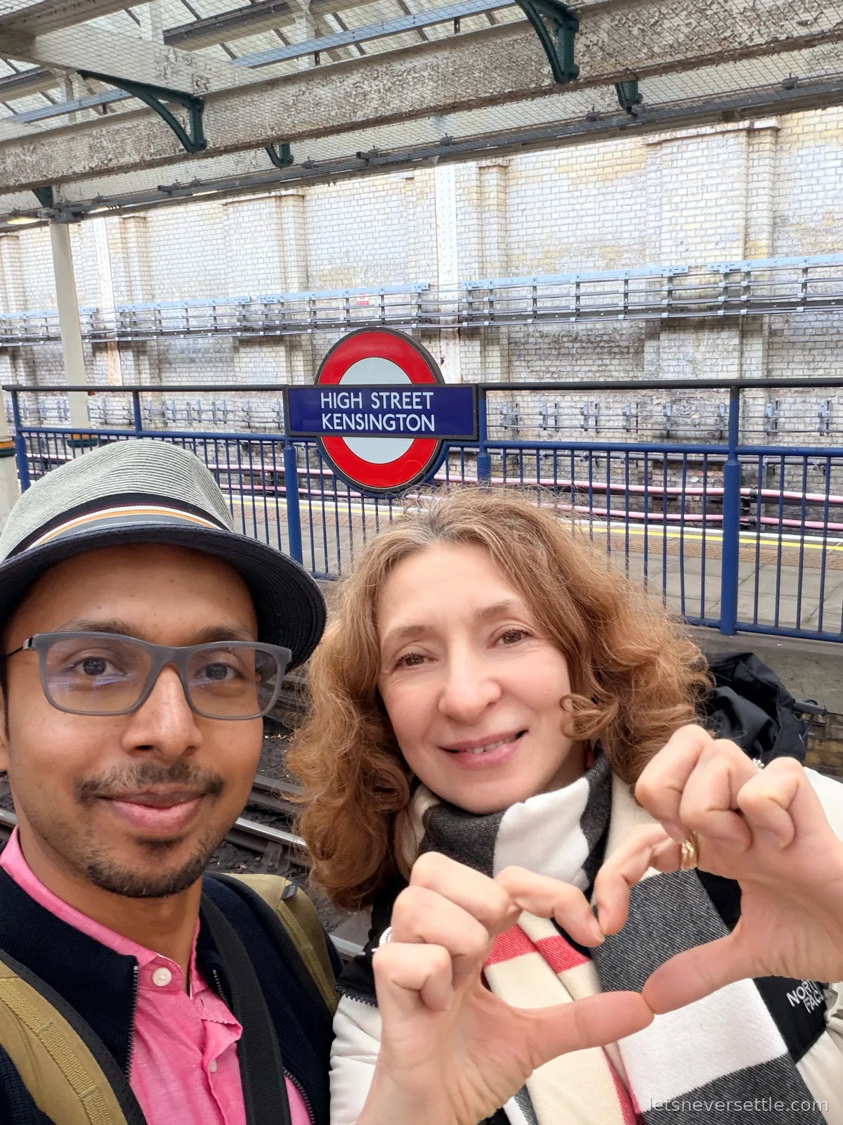 Selfie of Dileep in a trilby and glasses with Inga in a check scarf making a shared heart with their hands on the platform at High Street Kensington station