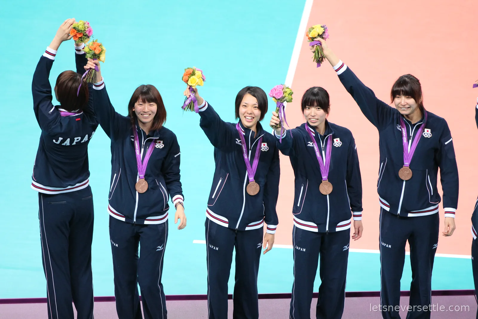 Japanese volleyball players in navy tracksuits waving flowers with bronze medals around their necks at London 2012
