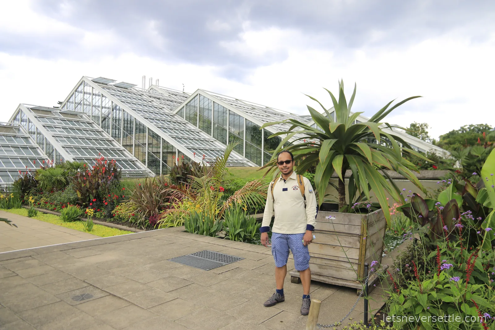 Dileep standing beside a tall aloe in a wooden planter outside the angular sloping glass roofs of the Princess of Wales Conservatory at Kew