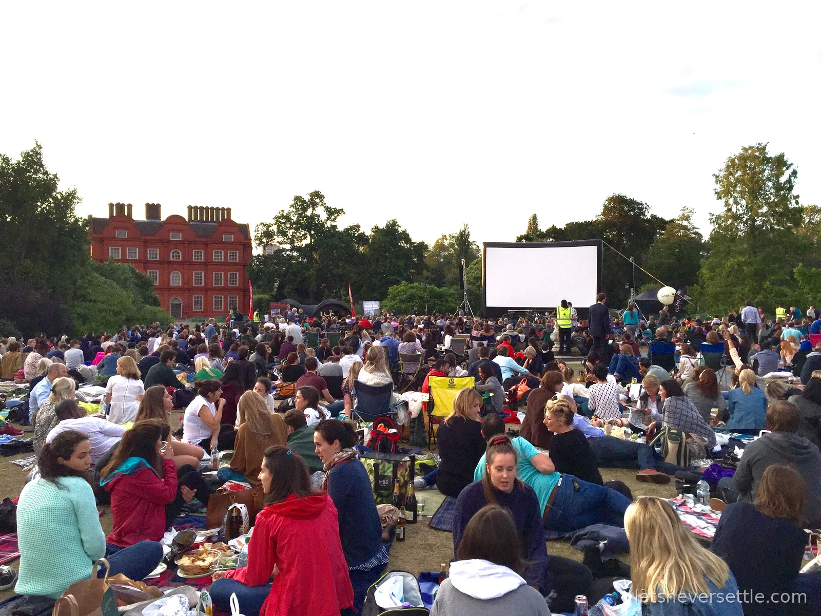 A large open air cinema screen on the lawn at Kew Gardens with hundreds of people sitting on picnic blankets, the red brick front of Kew Palace visible through the trees