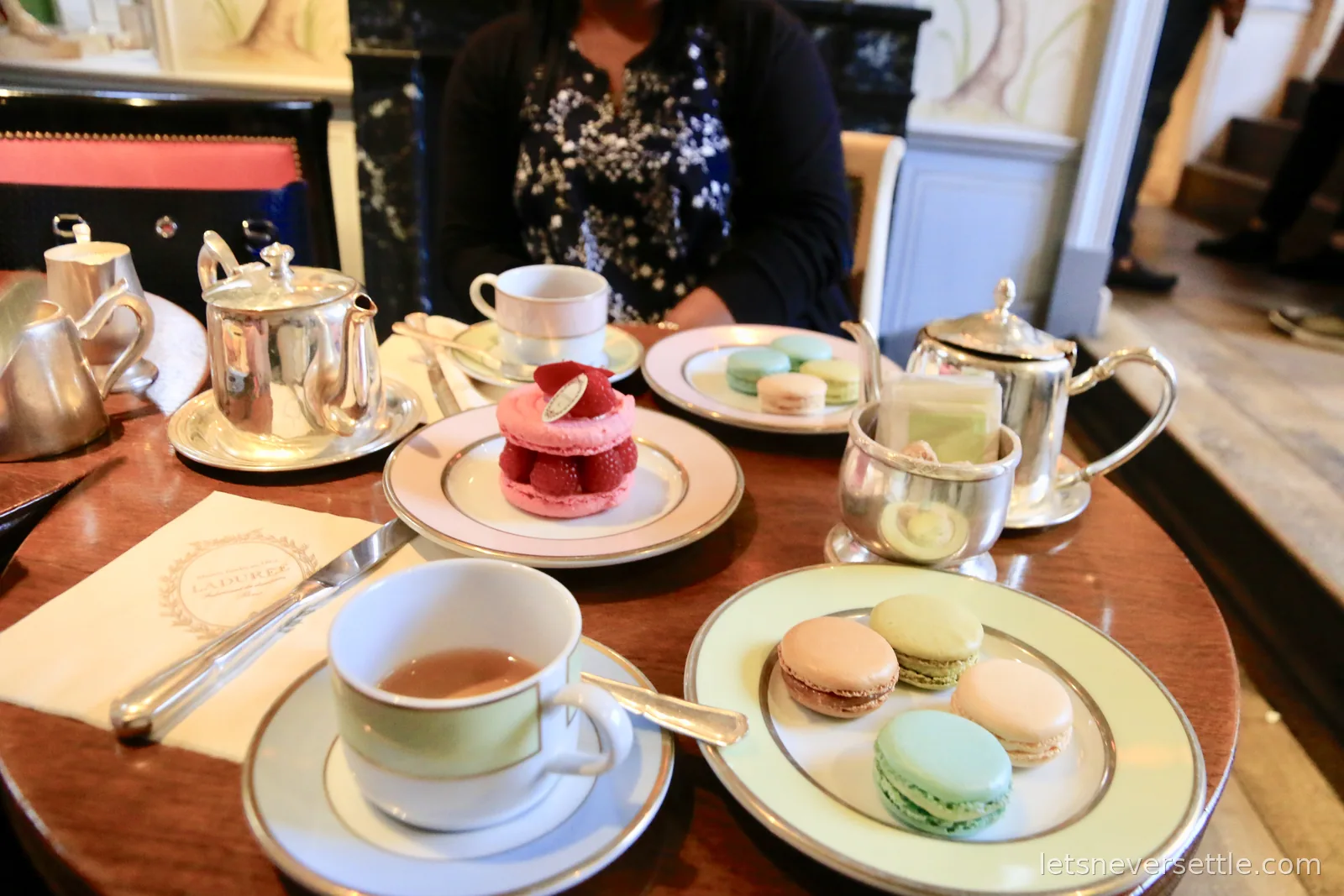Plates of pastel macarons, a large pink macaron topped with fresh raspberries, and silver teapots on a wooden table at Laduree