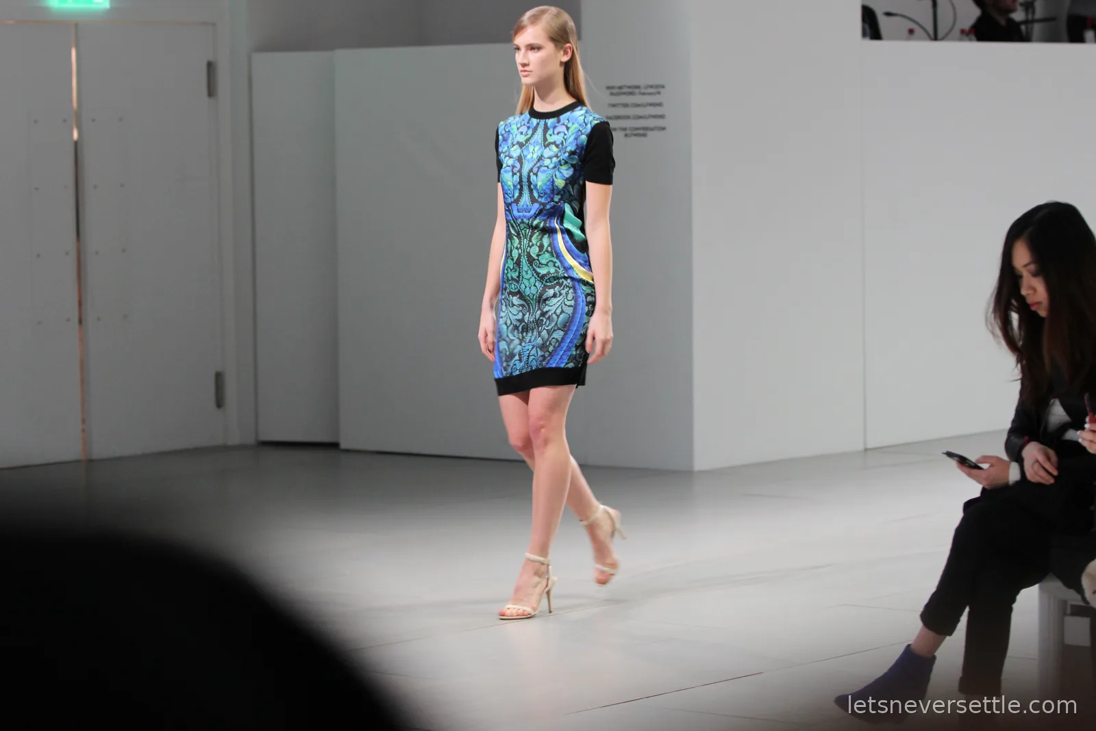 A model on the catwalk in a blue and green printed dress with black sleeves and nude heels, a spectator checking her phone at the edge of the runway