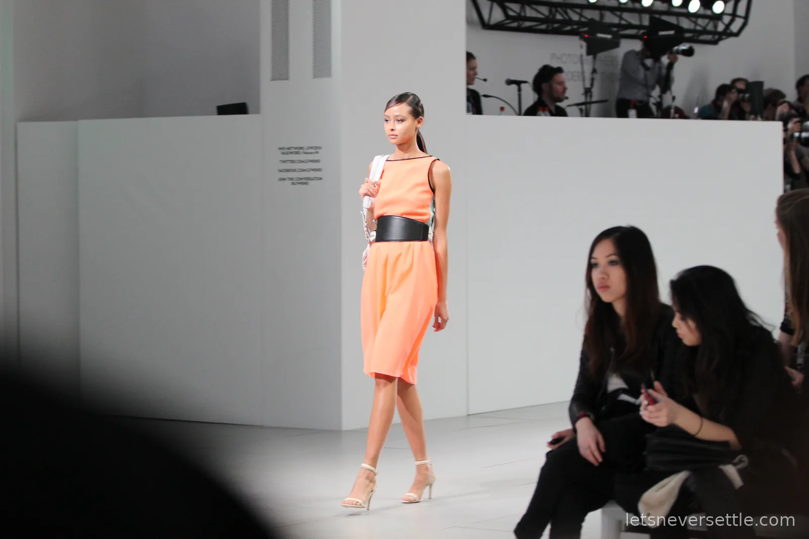 A model in a sleeveless coral dress with a wide black waist belt and white heels, audience members seated along the runway