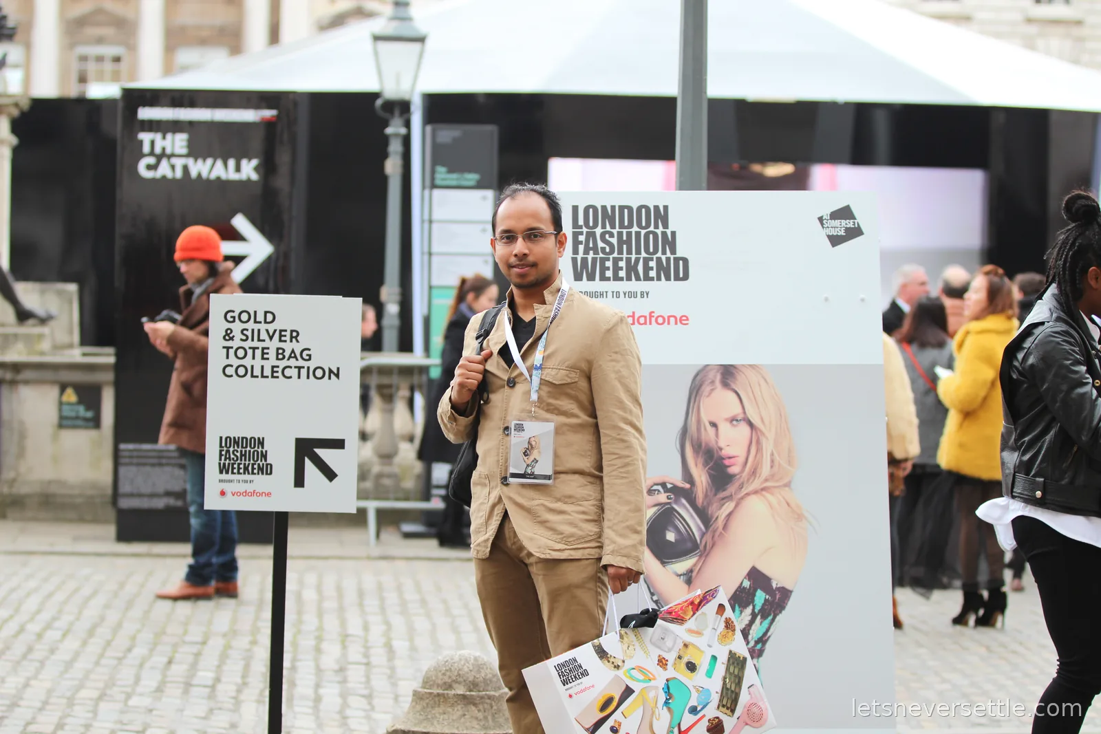 Dileep with a lanyard and tote bag in the cobbled courtyard of Somerset House, London Fashion Weekend boards and a sign for the gold and silver tote bag collection around him