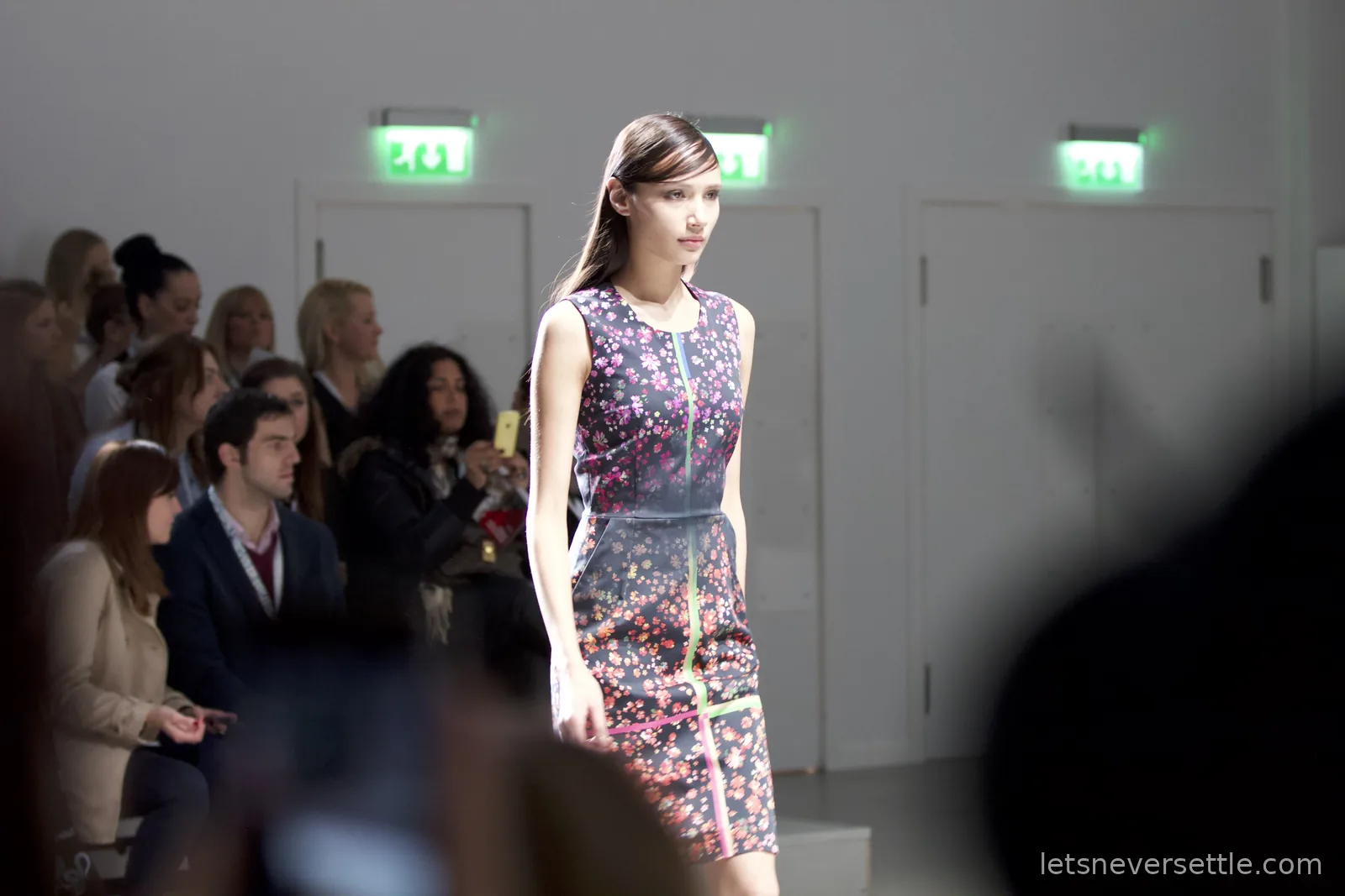 A model in a dark sleeveless dress scattered with pink and orange flowers walking past a seated audience