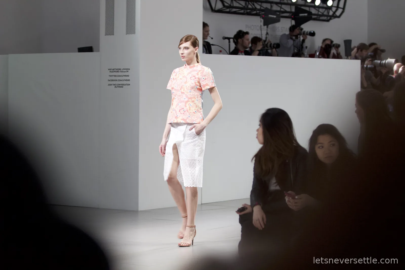 A model in a peach floral top and white broderie anglaise skirt with a front split, photographers massed at the end of the runway