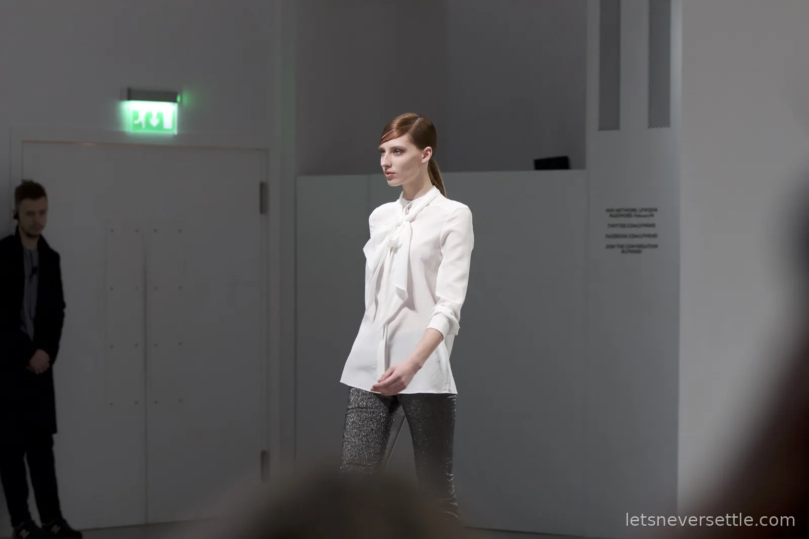 A model in a white pussy-bow blouse and sparkly silver trousers on the catwalk in front of white double doors