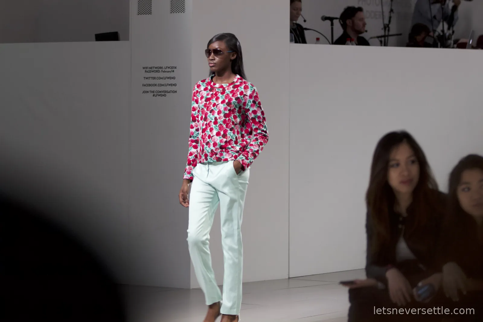 A model in sunglasses wearing a red rose print cardigan and mint green trousers, hands in pockets on the catwalk