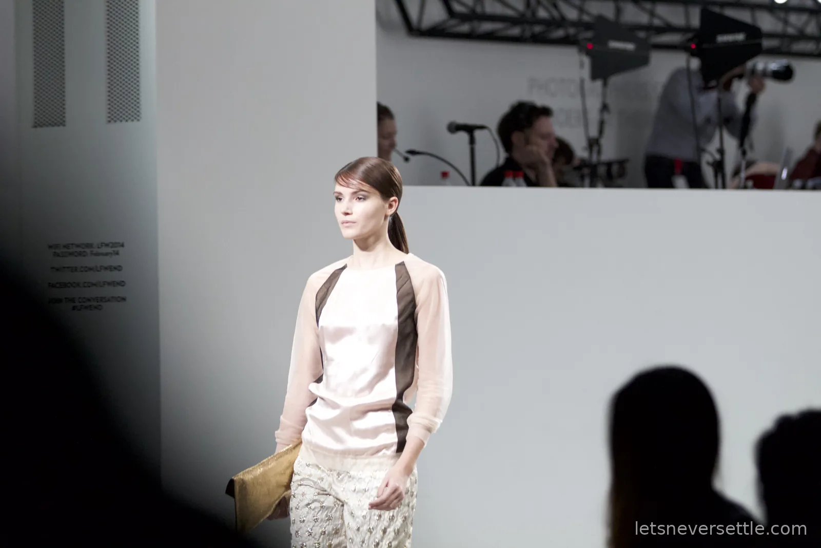 A model in a pale pink top with sheer sleeves and dark panels carrying a gold clutch, the photographers' stand visible behind the white catwalk wall