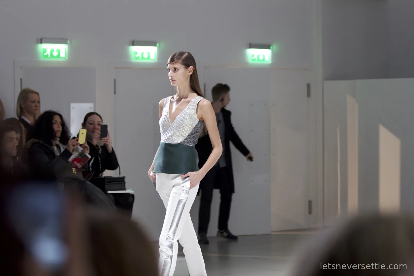 A model in a silver v-neck top with a dark green waistband and white trousers with a silver stripe, audience members filming on phones from the front row