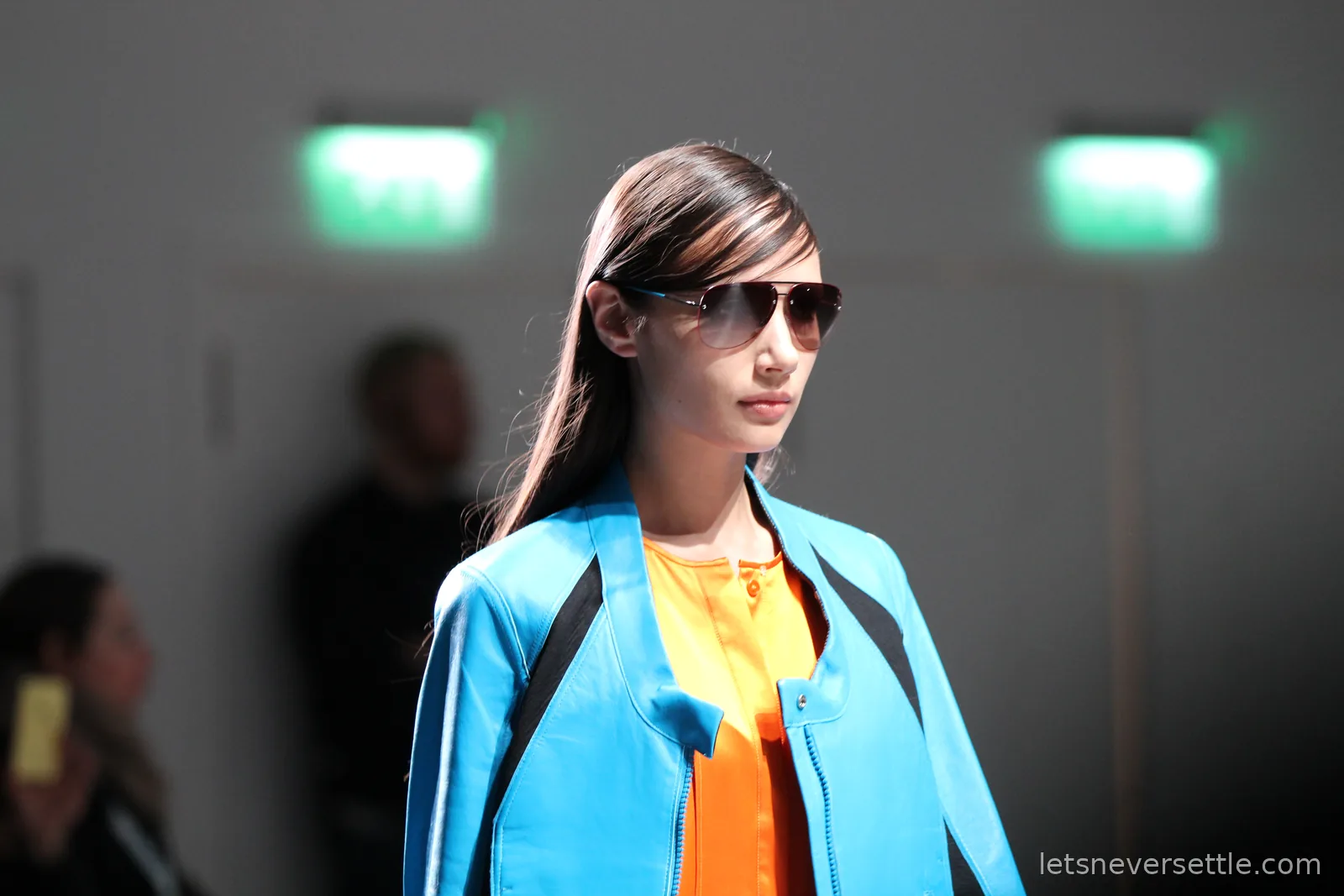 Close-up of a model in gradient aviator sunglasses, a bright turquoise leather jacket open over an orange shirt