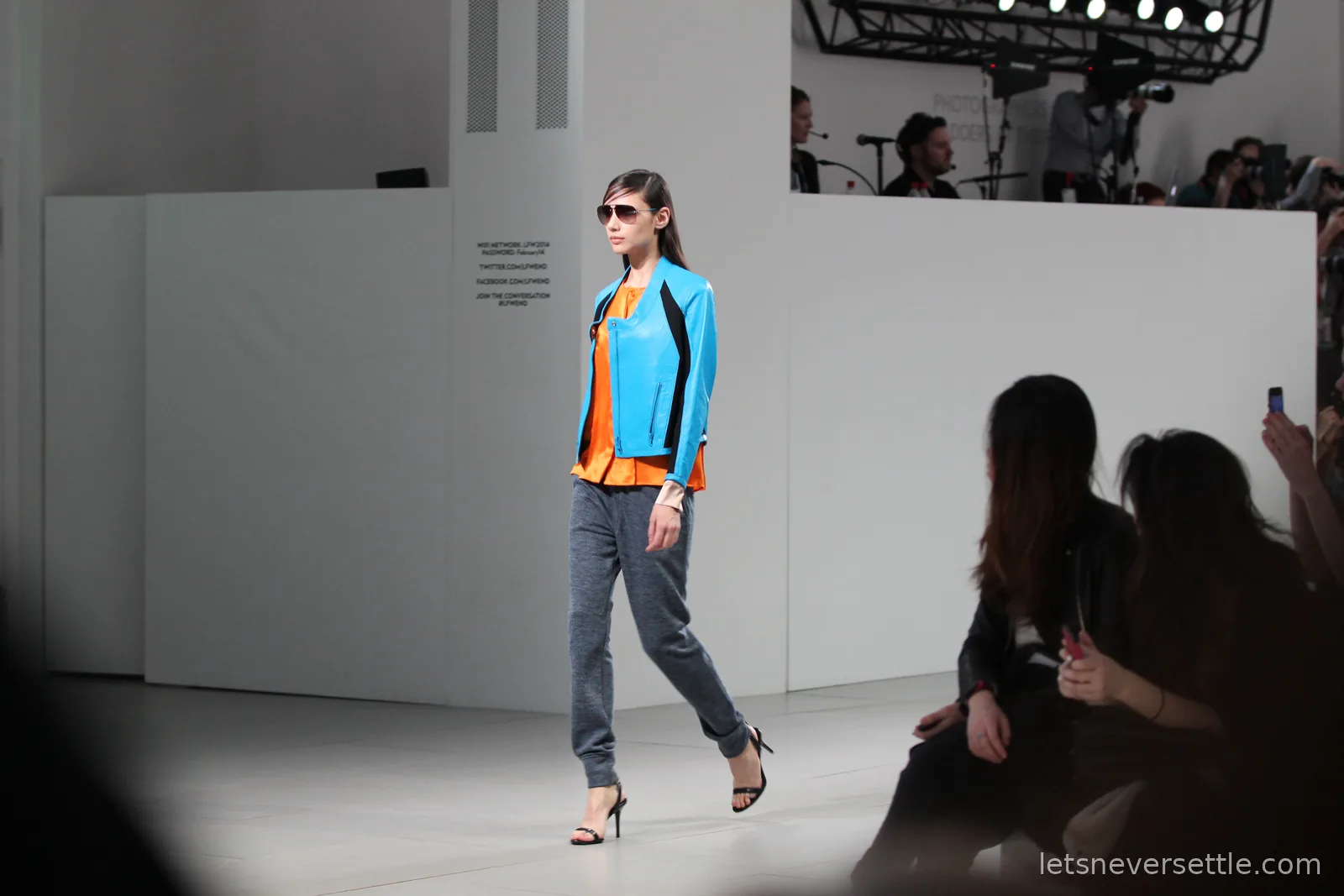 A model in sunglasses wearing a turquoise leather jacket over an orange top with grey jogger trousers and black heels on the catwalk