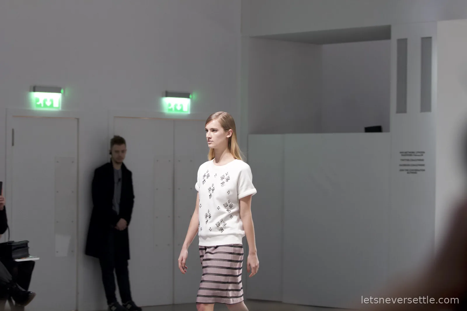 A model in an embellished white short-sleeved jumper and striped pencil skirt walking the white catwalk past a security guard