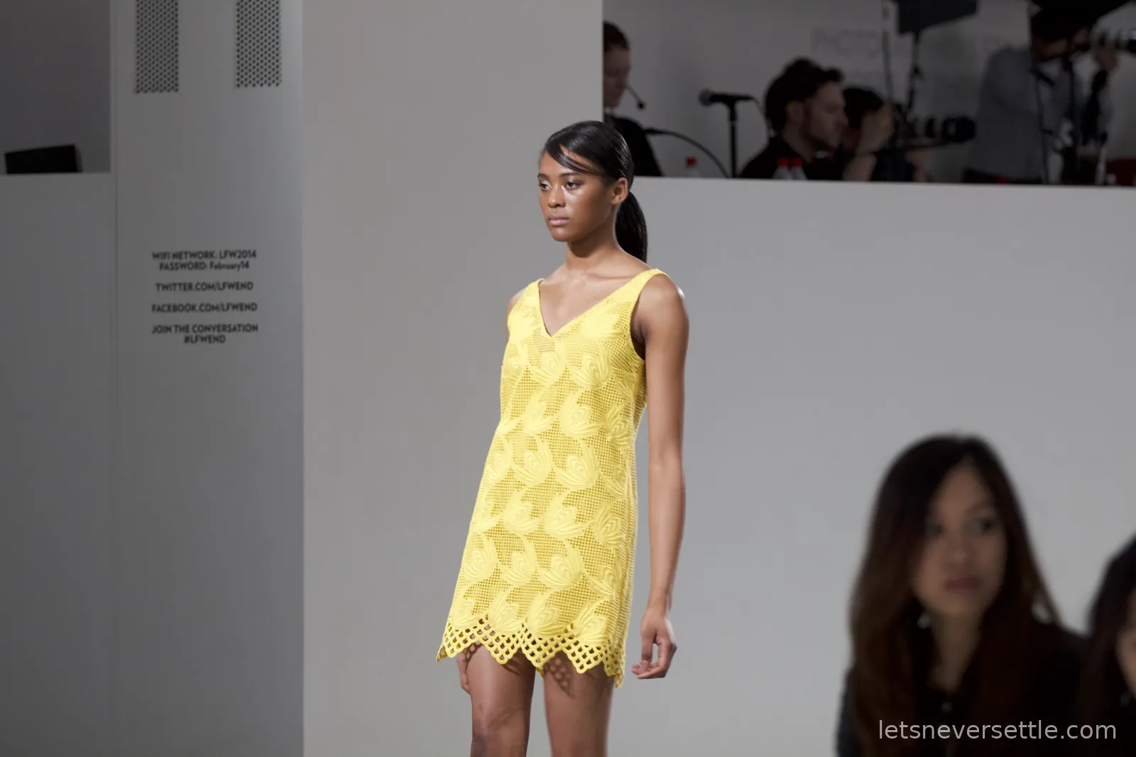 A model in a yellow lace shift dress with a scalloped hem on the catwalk, the venue wifi details printed on the wall beside her