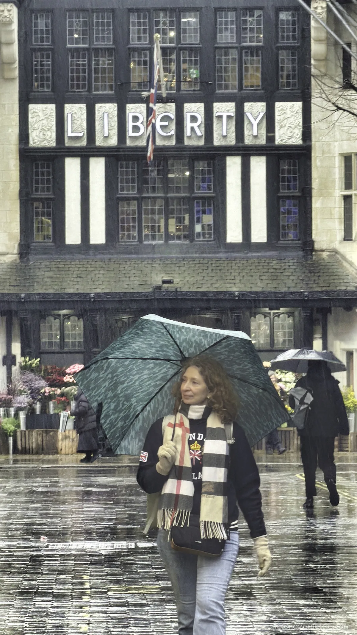 Inga under a teal umbrella walking past the black and white mock Tudor front of Liberty in heavy rain, the LIBERTY lettering and a Union Jack above her