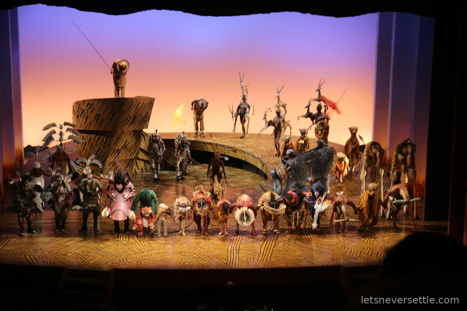 The full company of The Lion King on stage in animal costumes and masks gathered around Pride Rock against an orange sunrise backdrop